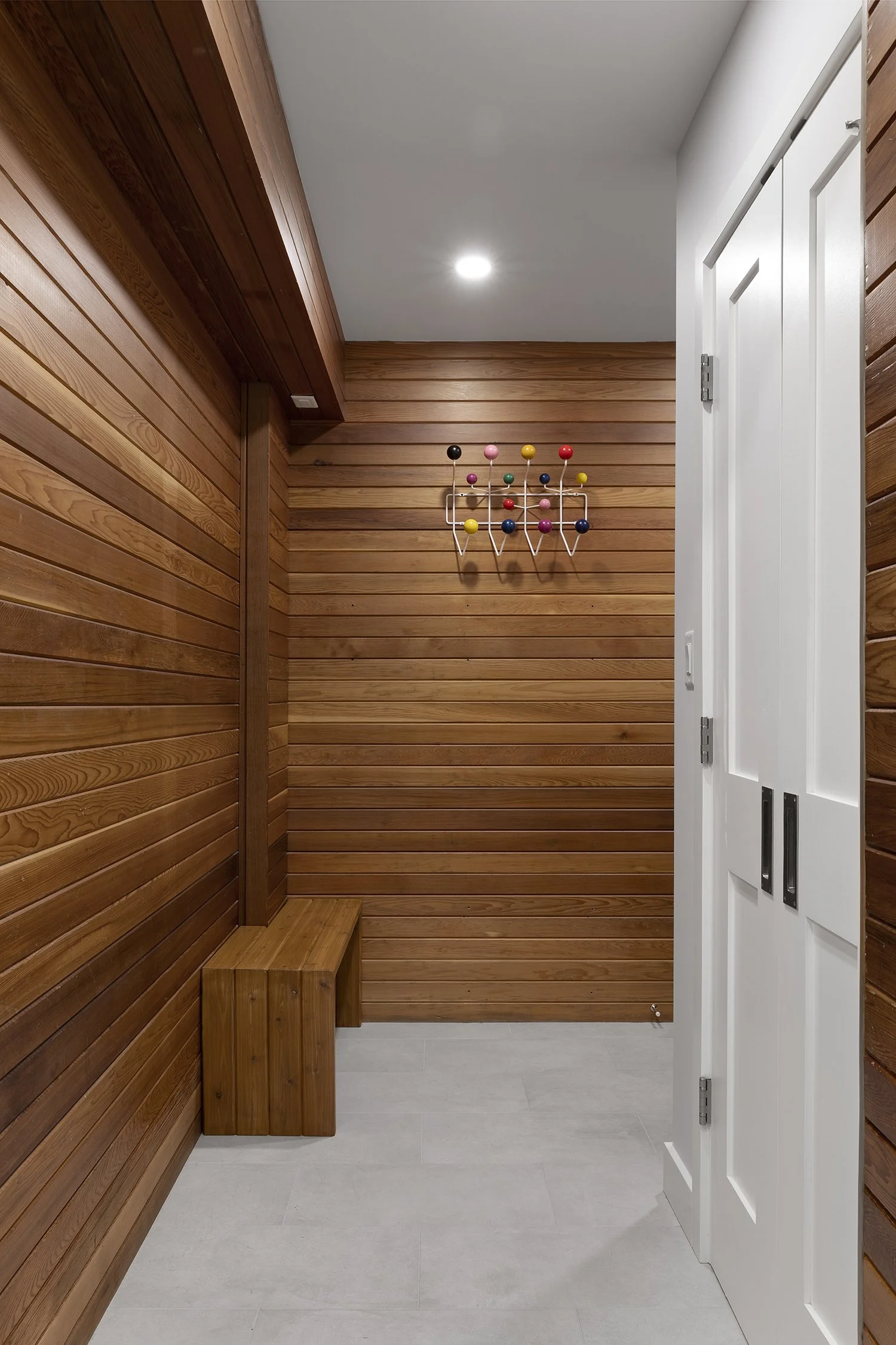 Wood-paneled walls with a white door open to reveal a small, minimalist interior space with a wooden bench and colorful wall-mounted amusement park-style game hook.