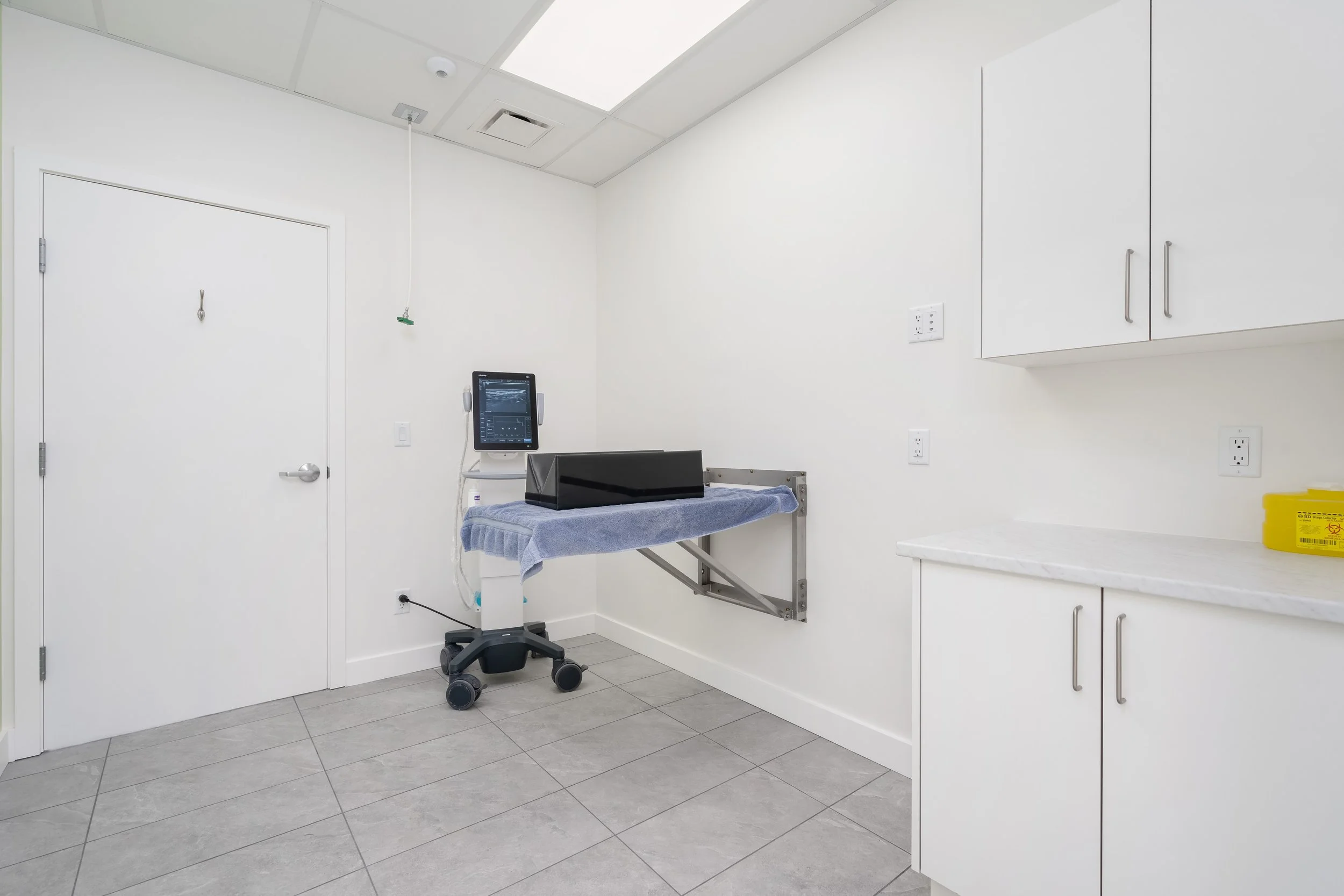 Empty hospital room with white walls, a closed white door, medical equipment including monitor, rolling stool, and a covered examination table with a blue towel.