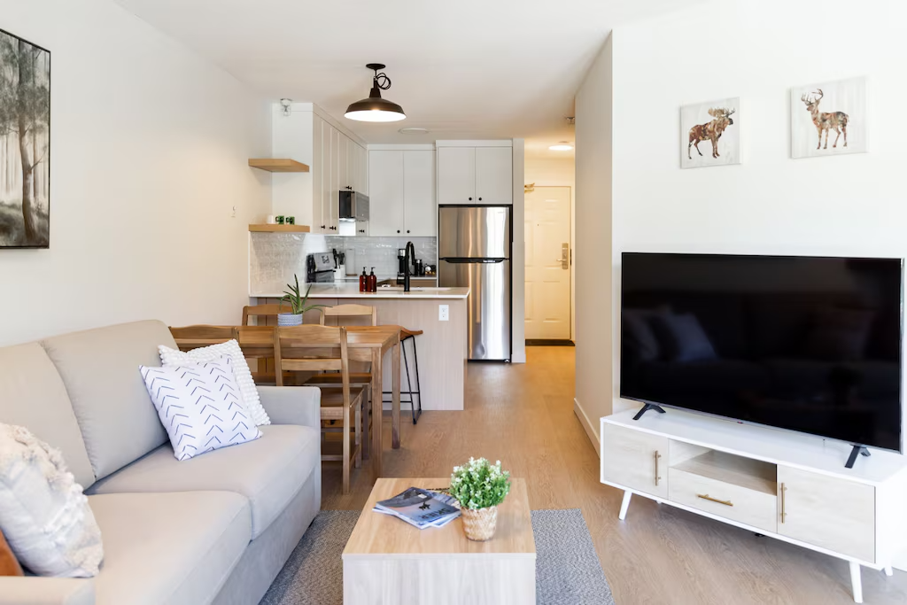 Open-concept living room and kitchen with beige sofa, wooden dining table, flat-screen TV on white stand, and kitchen cabinetry with stainless steel refrigerator.