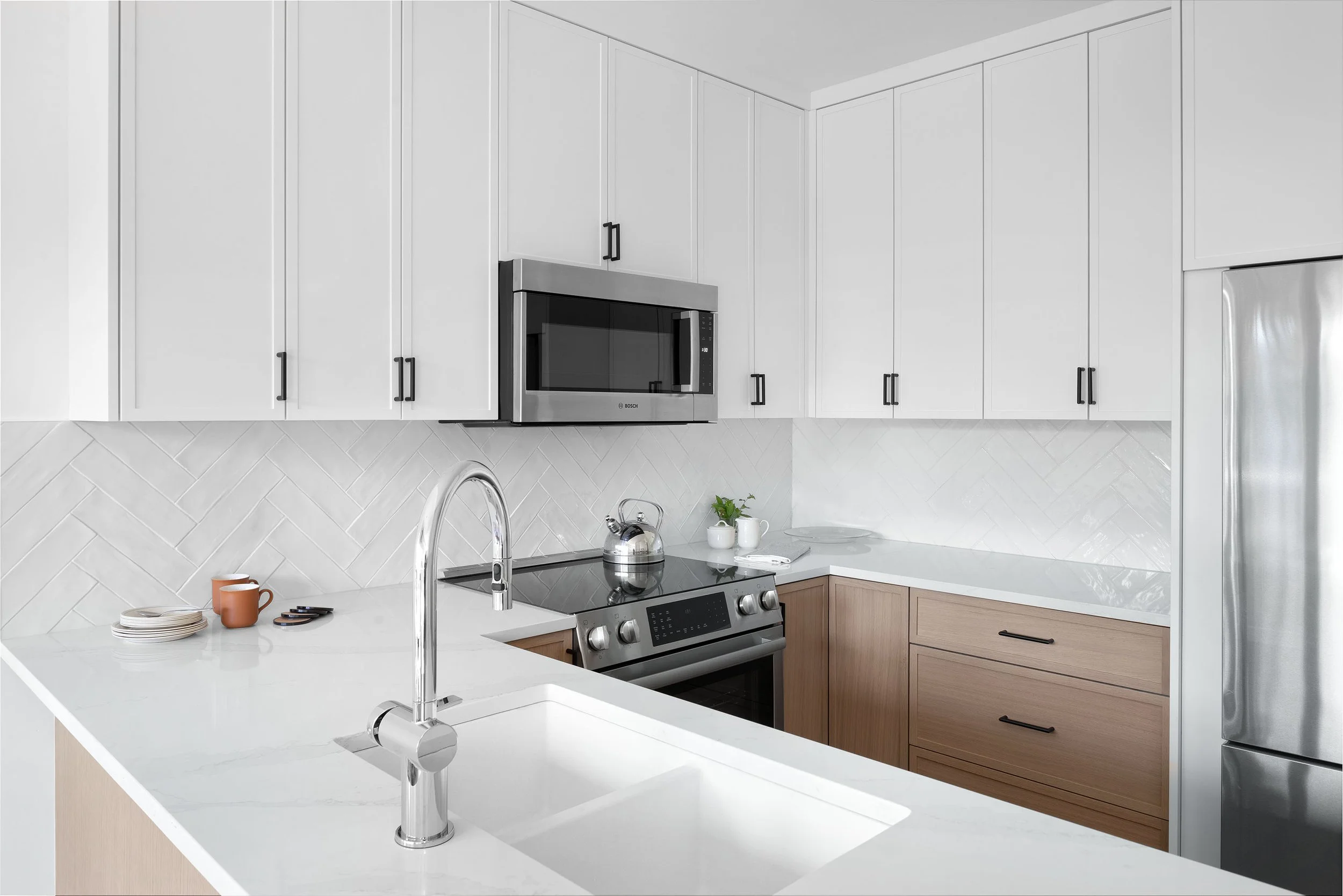 Modern kitchen with white cabinetry, stainless steel microwave and stove, white countertop, and light wood lower cabinets. A silver kettle and cups are on the counter, with a sink and faucet in the foreground.