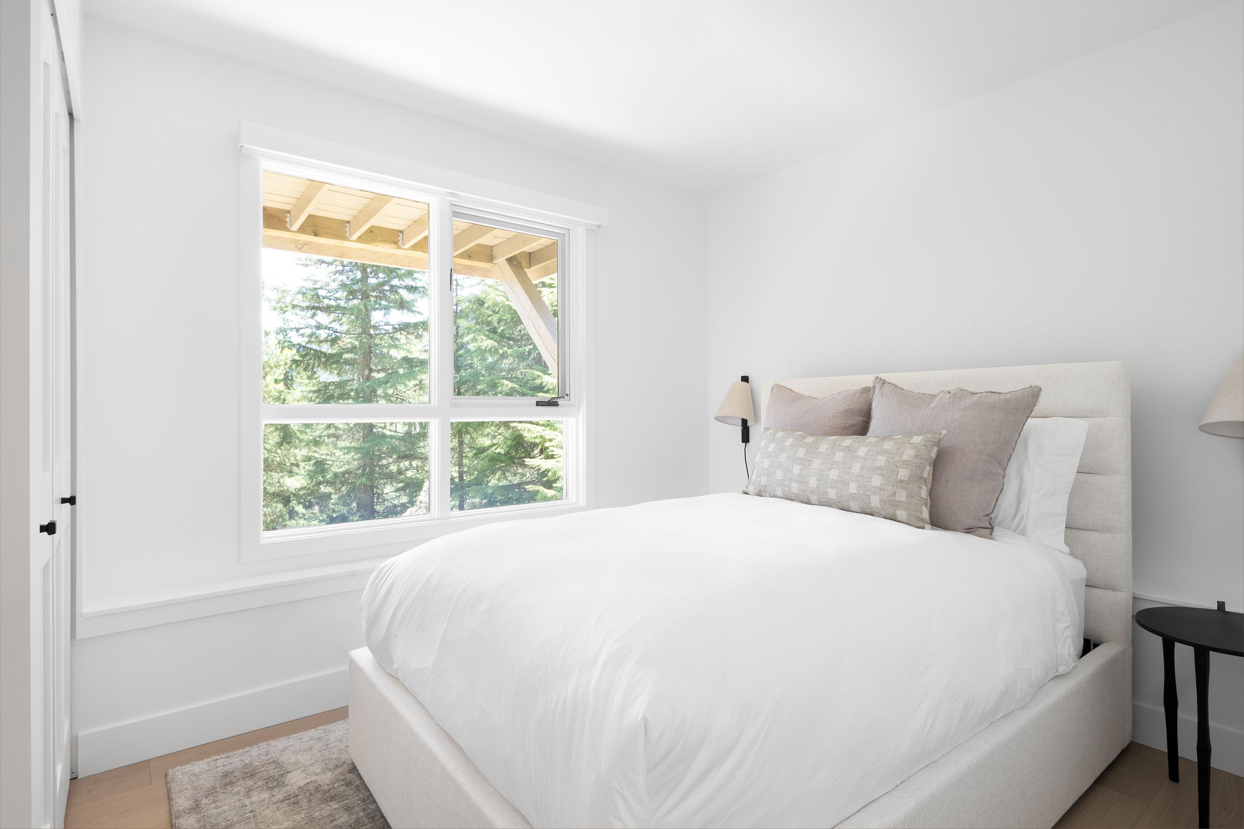A bedroom with a large window showing trees outside, a bed with white bedding and pillows, a beige headboard, two matching lamps on either side, and a small black side table.