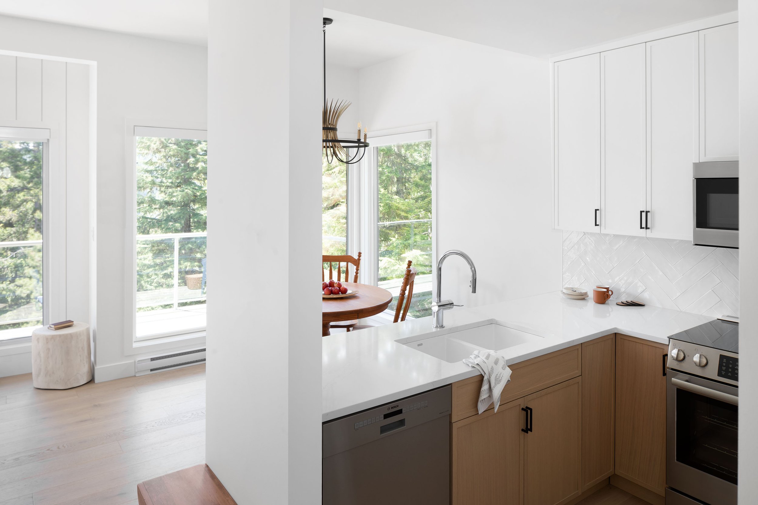 Modern kitchen with white walls, light wood cabinets, a white countertop, stainless steel appliances, and large windows showing trees outside.