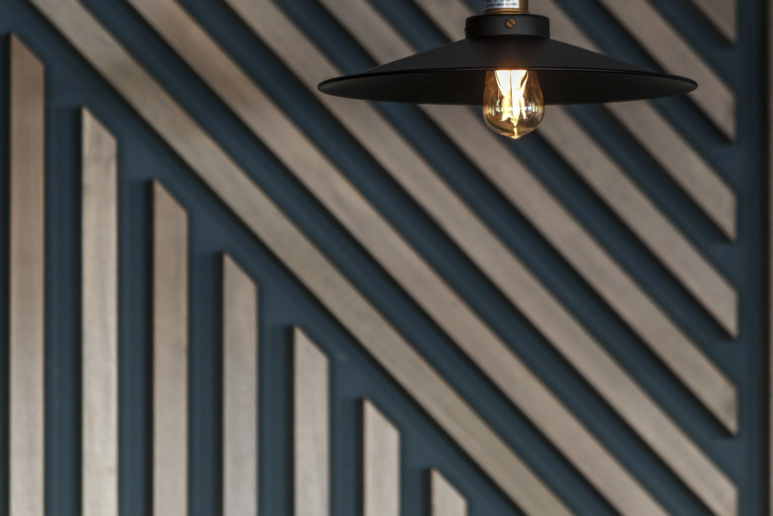 Close-up of a black pendant light with an exposed filament bulb hanging over a dark wooden slatted ceiling.