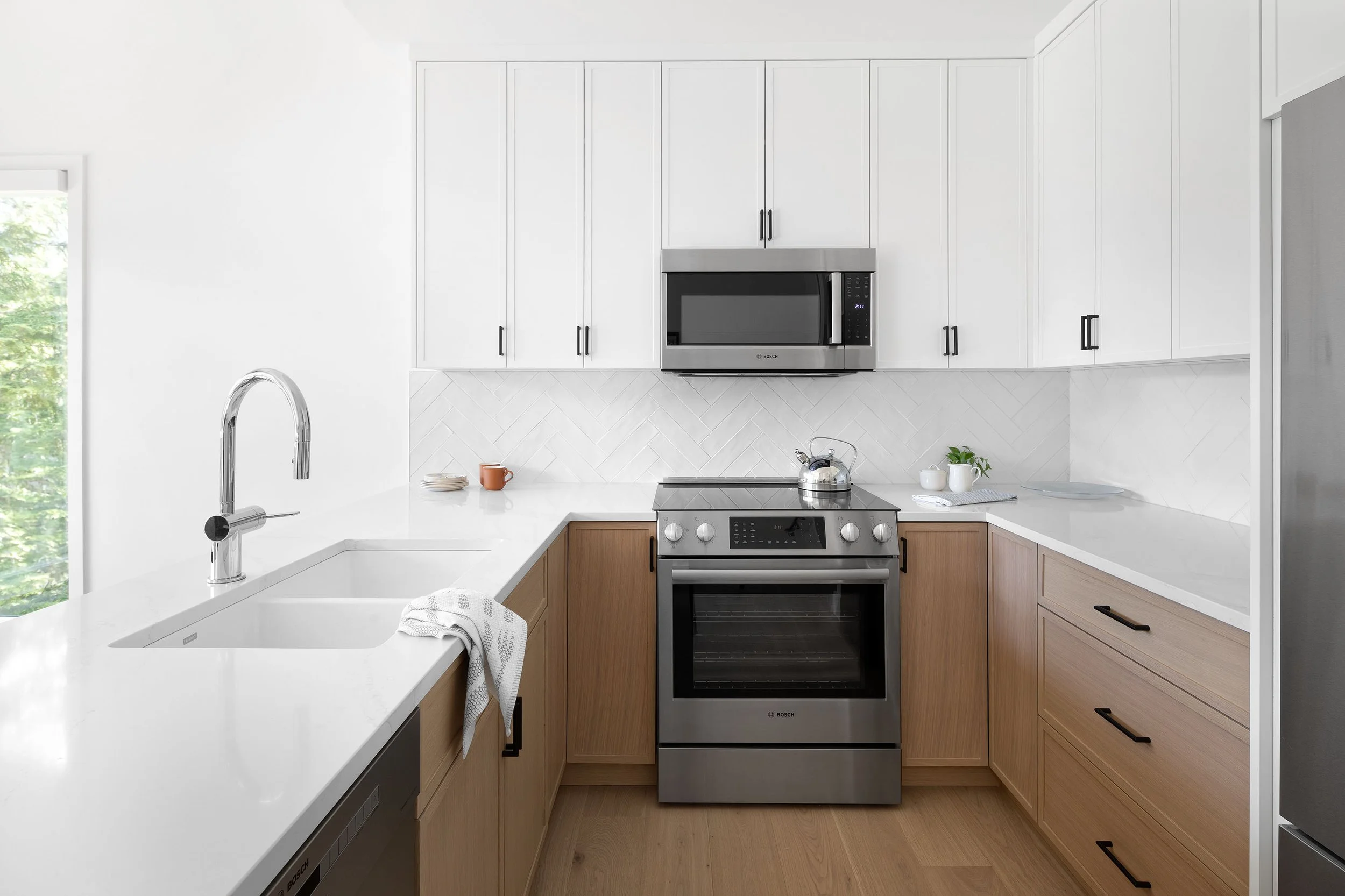 Modern kitchen with white upper cabinets, wood lower cabinets, stainless steel oven and microwave, white countertops, and a window with greenery outside.