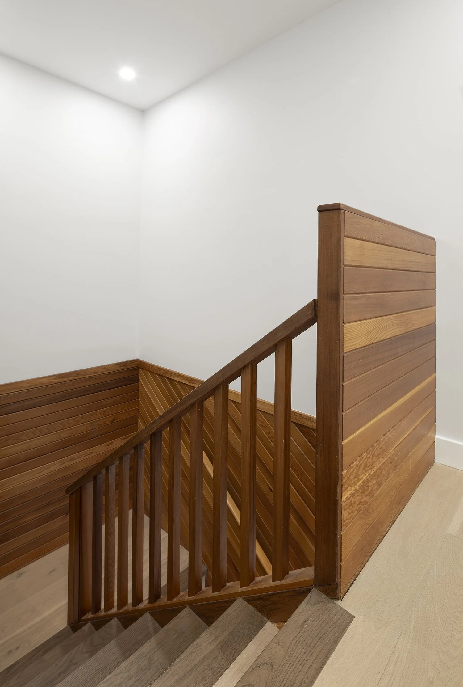 Wooden staircase with a wooden railing and wall paneling, leading down to a beige carpeted floor, in a minimalist interior with white walls and a ceiling light.