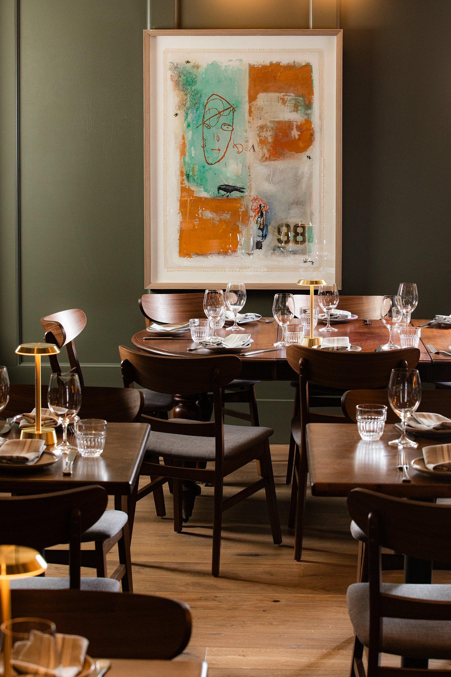 A dining room with several wooden tables and chairs, set with glasses, napkins, and utensils. A large abstract painting with orange, light green, gray, and black colors hangs on the green wall behind the tables. The room is softly lit with warm light