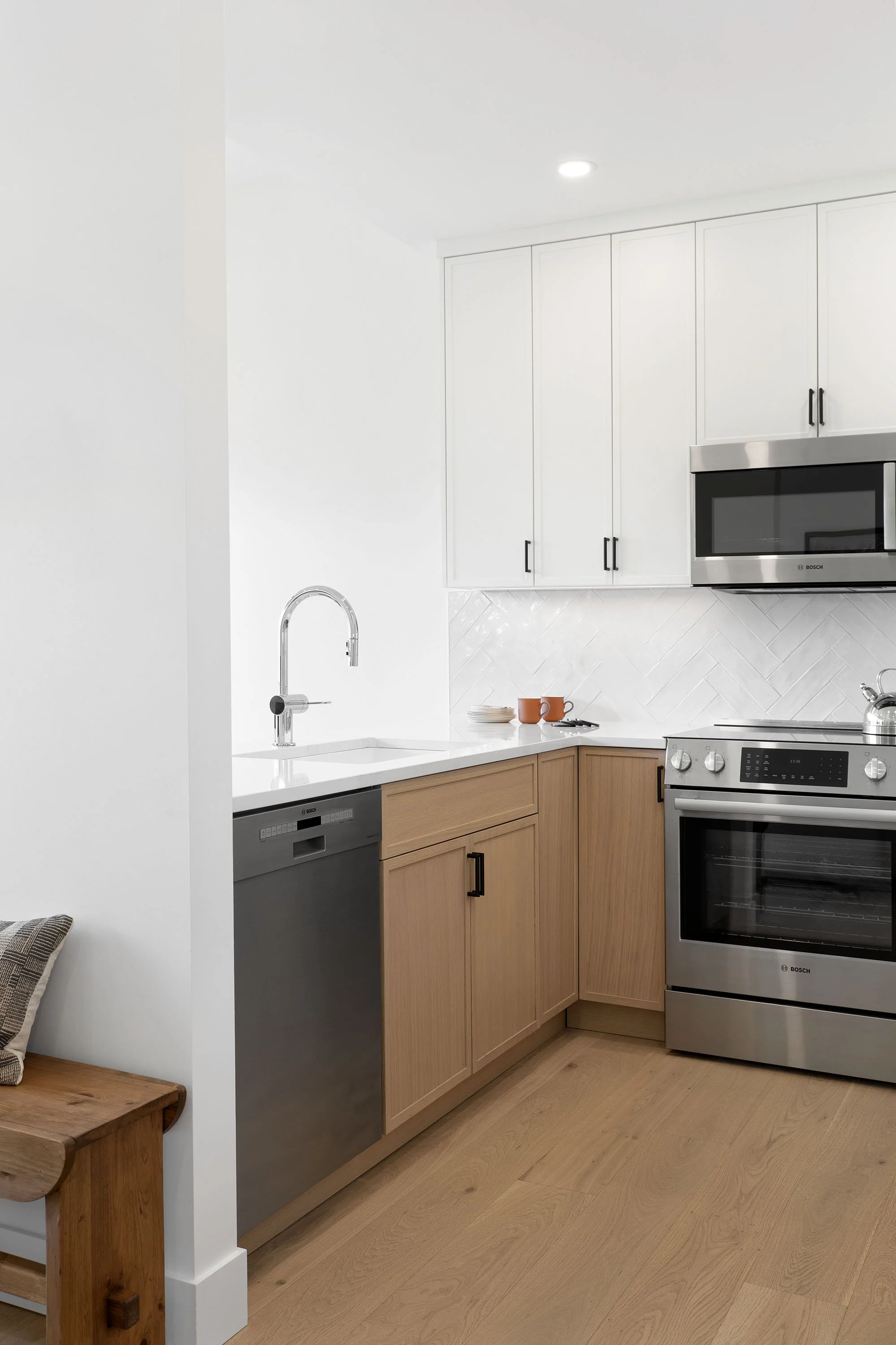 Modern kitchen with white cabinets, a stainless steel microwave, and a range, with light wood lower cabinets and flooring, and a small seating area with a plaid cushion.
