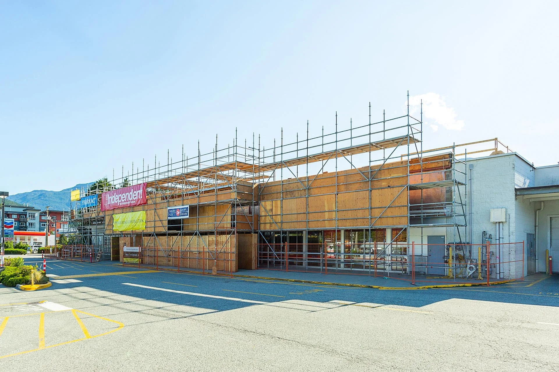 A building under construction with scaffolding around it. The building has wooden panels and is next to a white structure. There are banners and a parking lot in front, with mountains visible in the background and a clear blue sky.