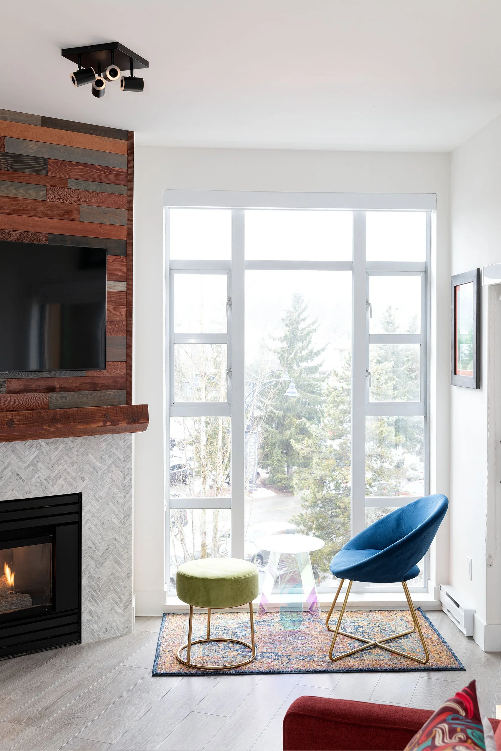 A well-lit living room with a large window, a green ottoman, a blue chair with gold legs, a colorful area rug, a wood and stone fireplace, and a wall with a wooden accent.