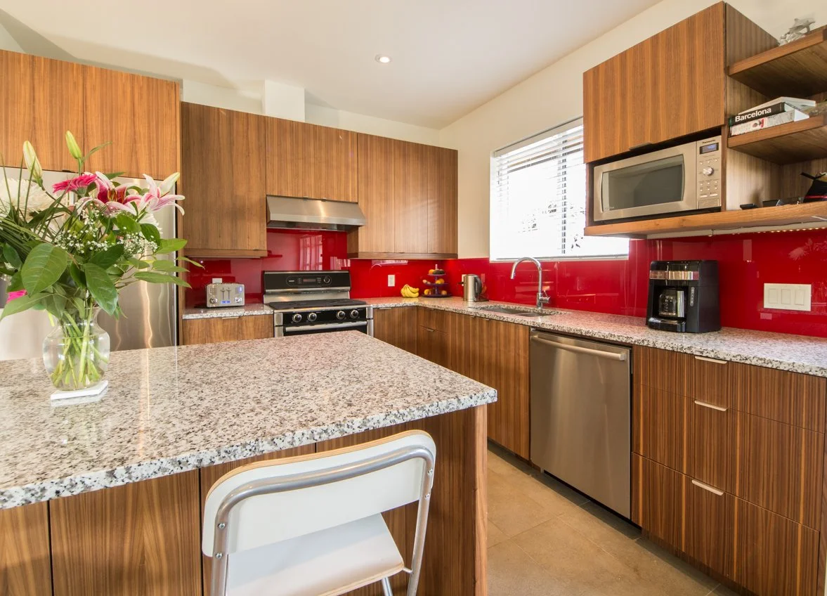 Modern kitchen with wooden cabinets, granite countertops, red backsplash, stainless steel appliances including a stove, dishwasher, and microwave. A fresh flower arrangement in a glass vase is on the counter.