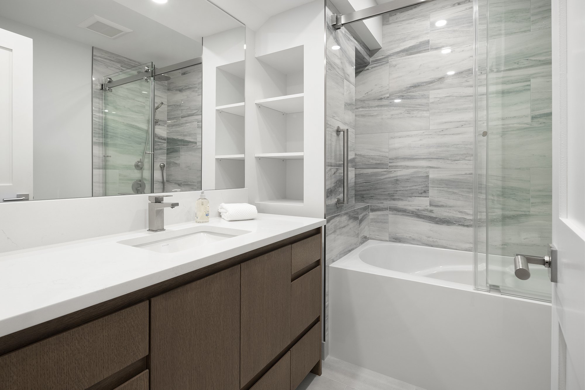 Modern bathroom with a white vanity, brown cabinetry, a sink, a large mirror, a soap dispenser, and a rolled white towel. There is a bathtub with a glass sliding door and a shower with gray marble tiles. White shelves are built into the wall above th