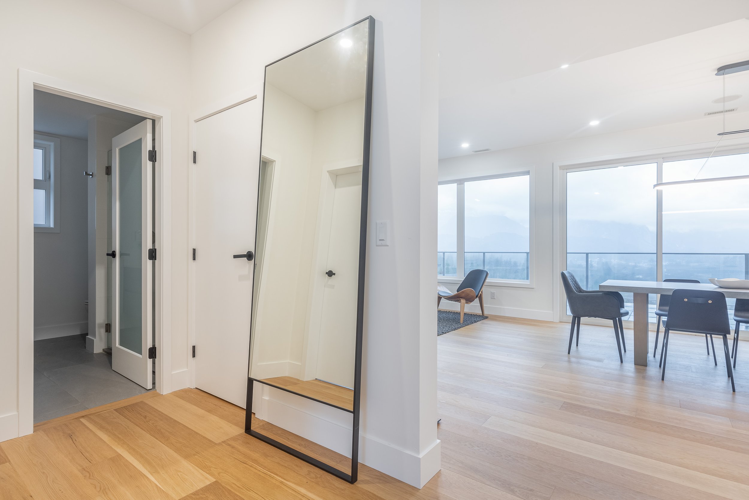 A modern, clean apartment interior with a large mirror, a dining table with four chairs, and large windows showing a city view.