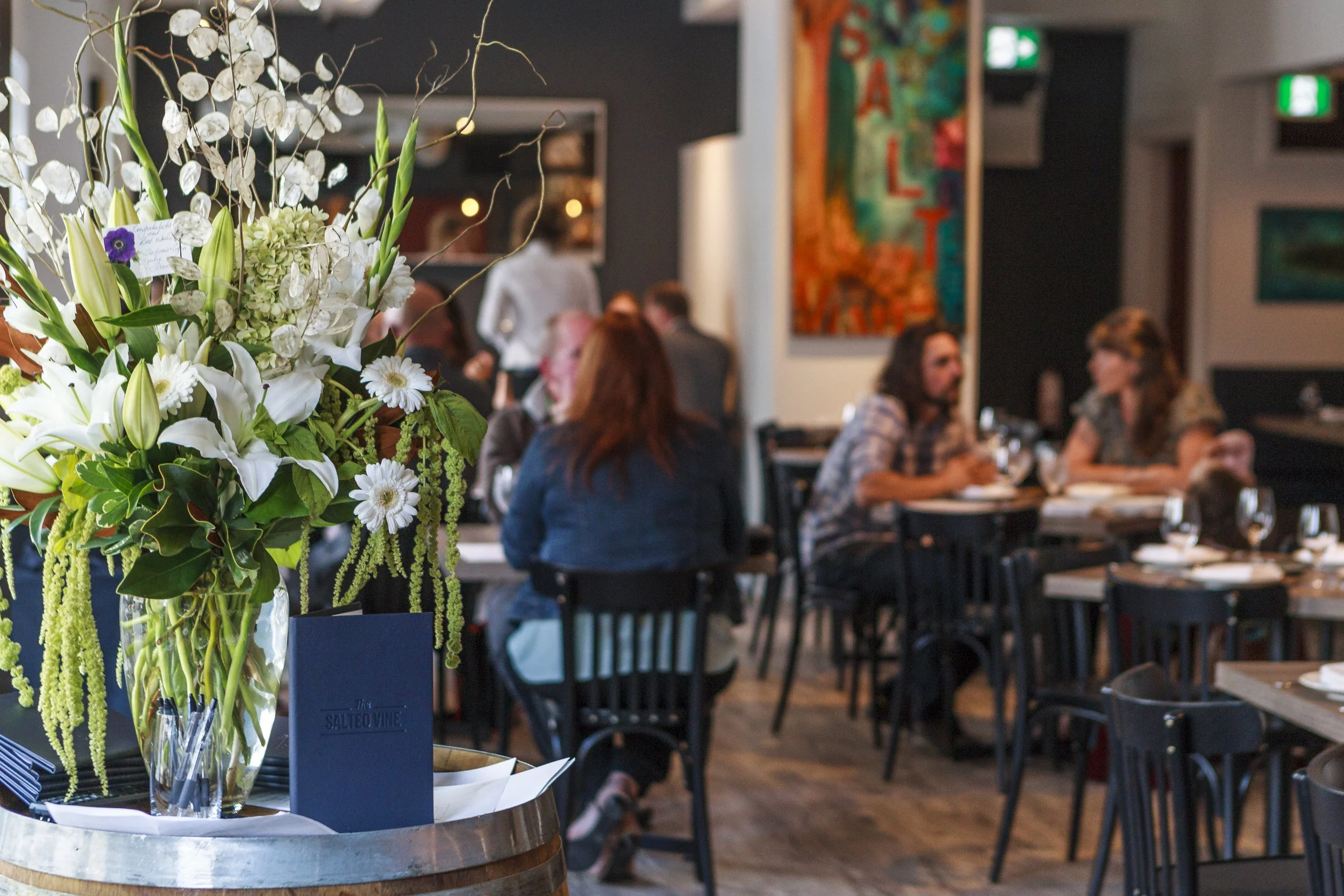 A floral centerpiece with white lilies, daisies, and other flowers on a table in a restaurant with people dining in the background.