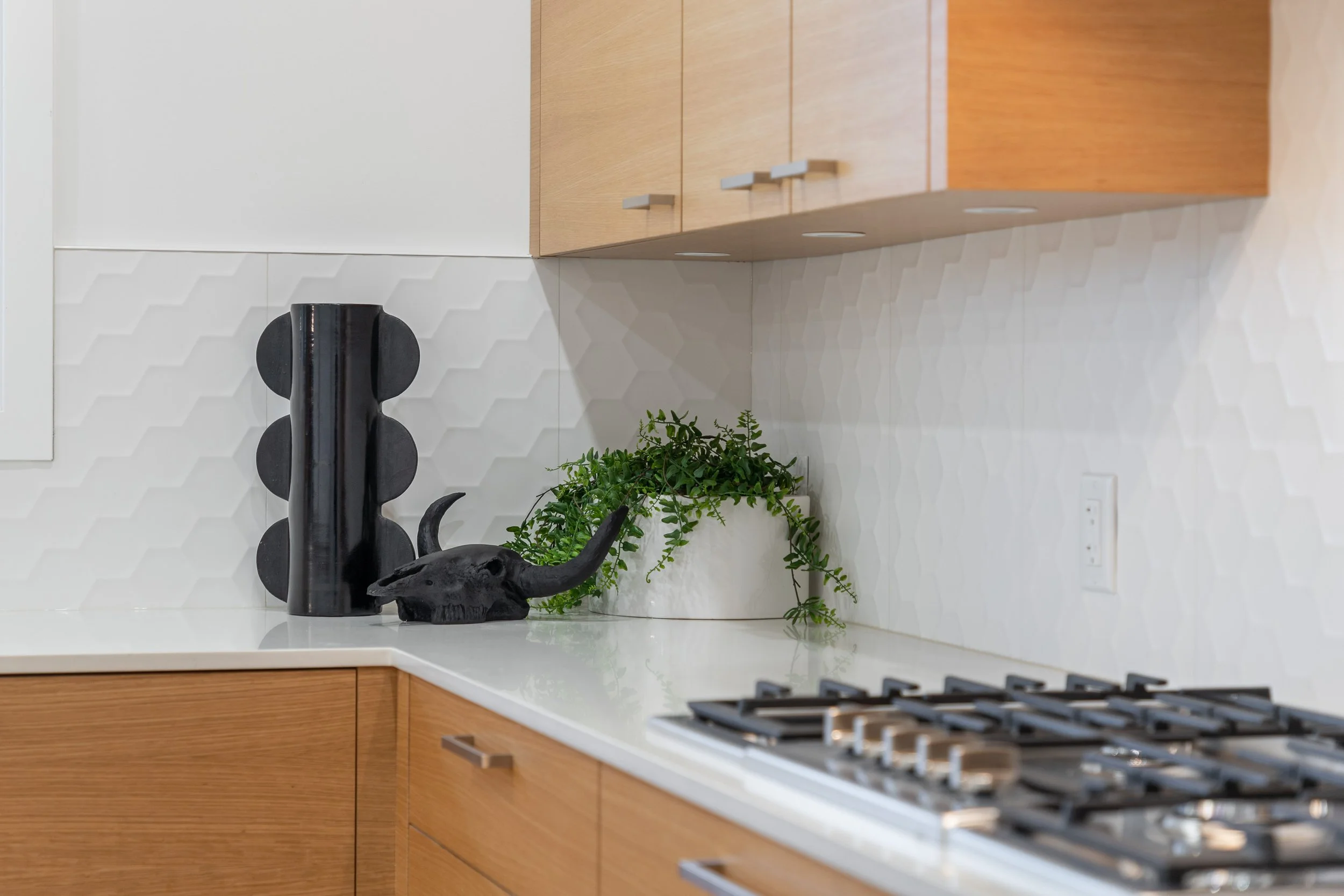 Kitchen countertop with a black decorative object resembling an animal skull, a green potted plant, and a black vase, with wooden cabinets and white hexagonal tile backsplash.