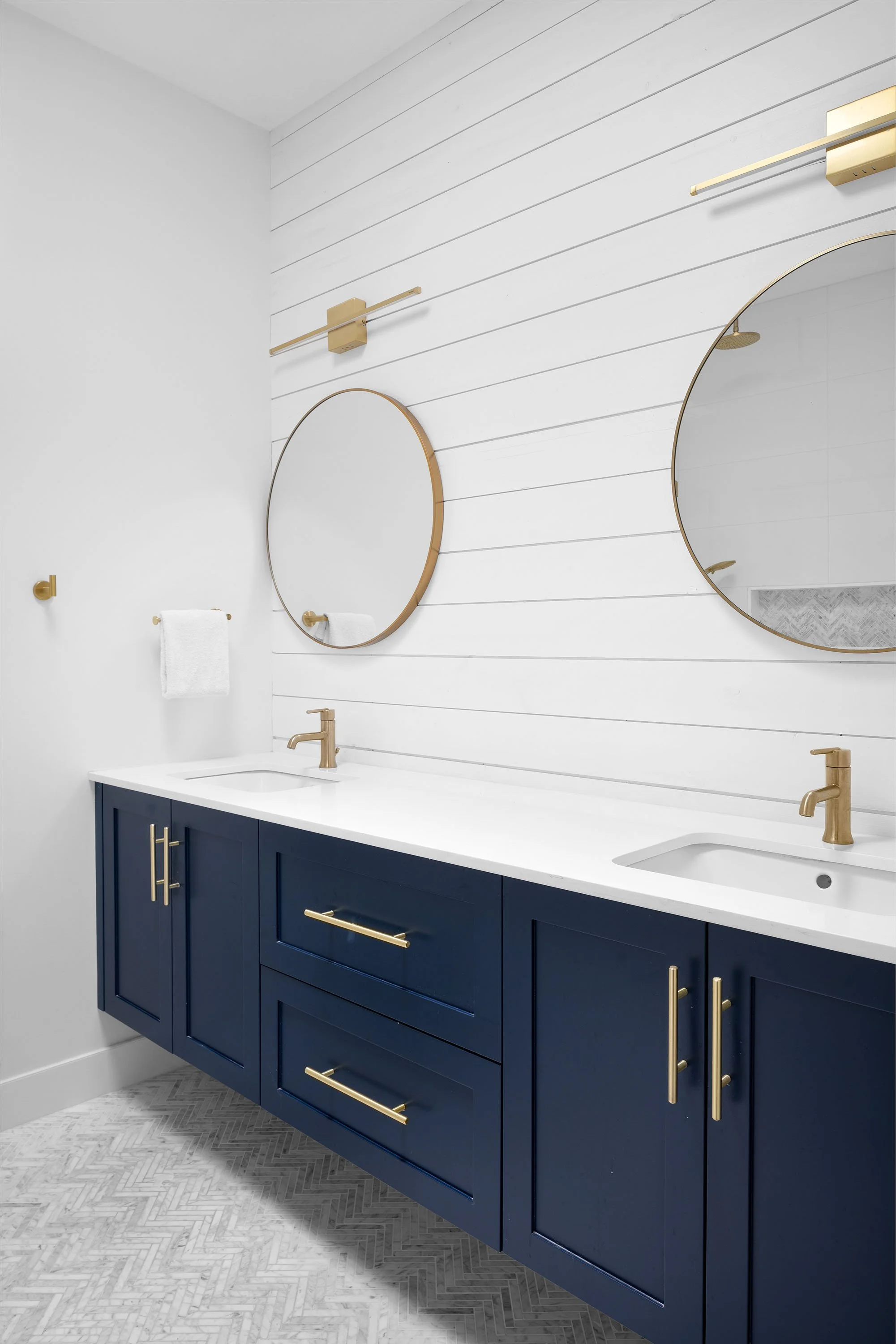 Bathroom vanity with a navy blue cabinet, white countertop, and gold hardware. Two round mirrors with gold frames hang on a white shiplap wall, with a towel rack and wall-mounted light fixtures above.