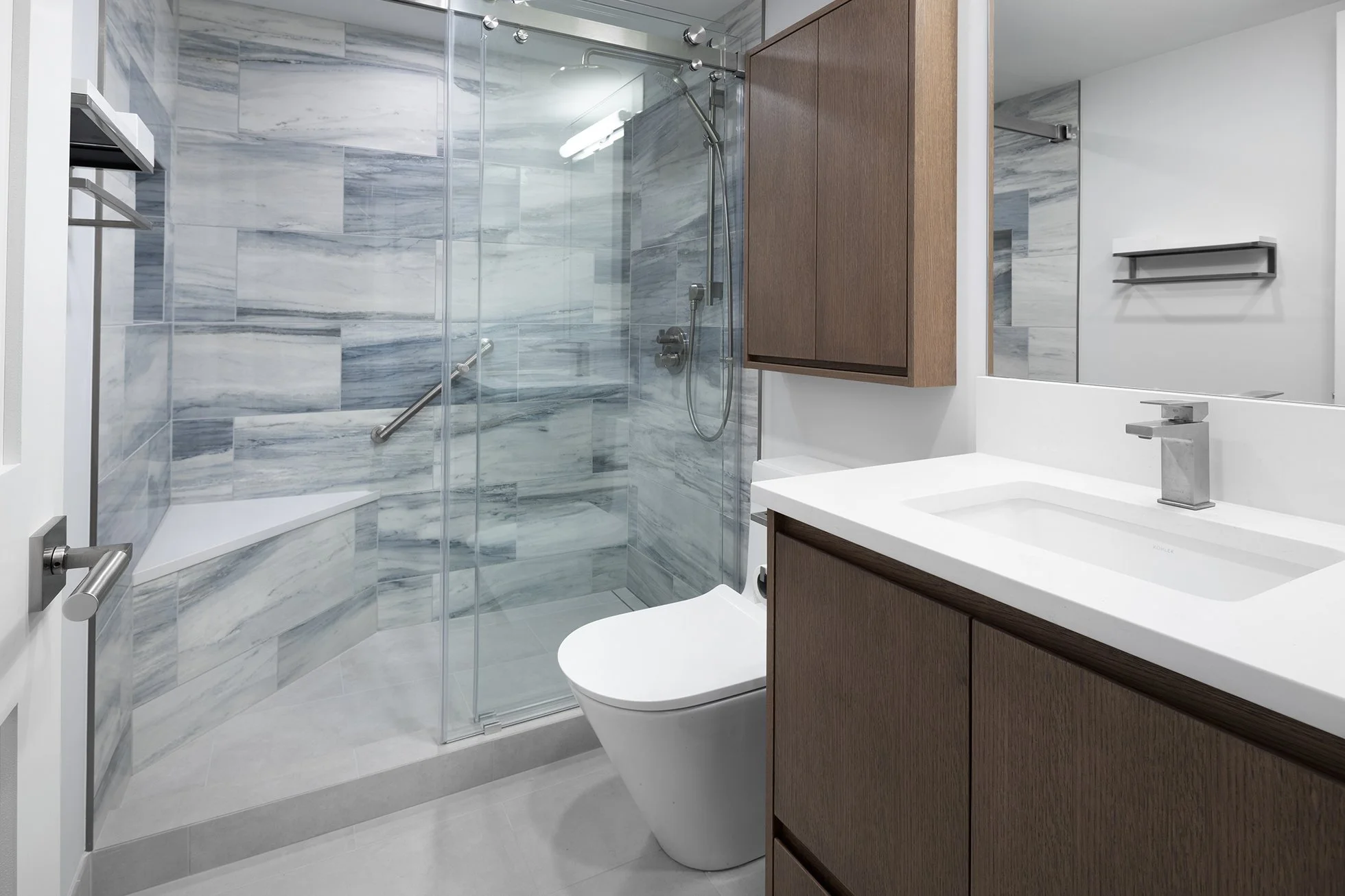 Modern bathroom featuring a walk-in shower with glass door and marble tile walls, a white toilet, a white countertop with integrated sink, and wooden cabinets.