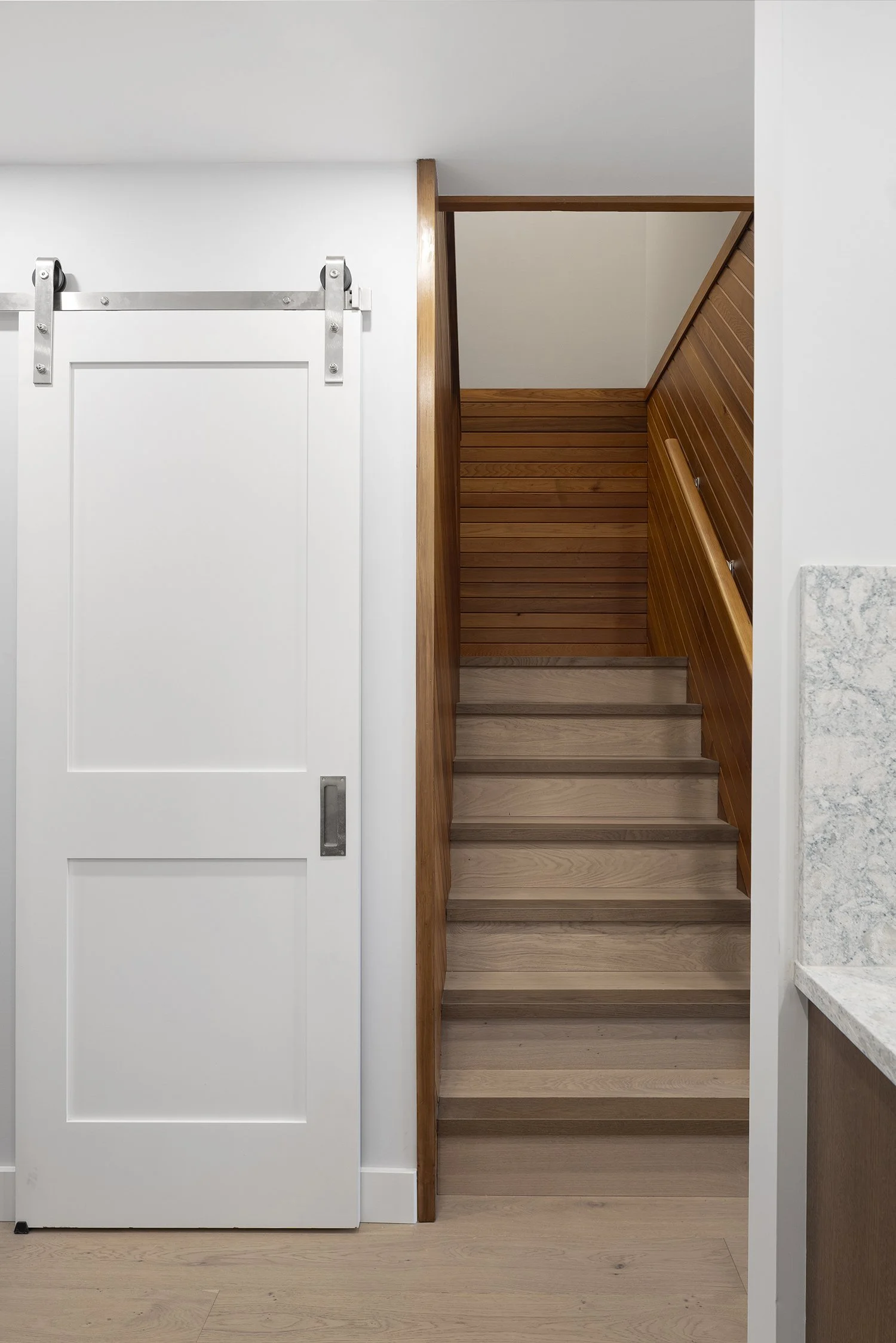 Interior view of a small home with a white sliding barn door on the left, a wooden staircase with a sloped wall on the right, and a countertop with a marble pattern on the far right.