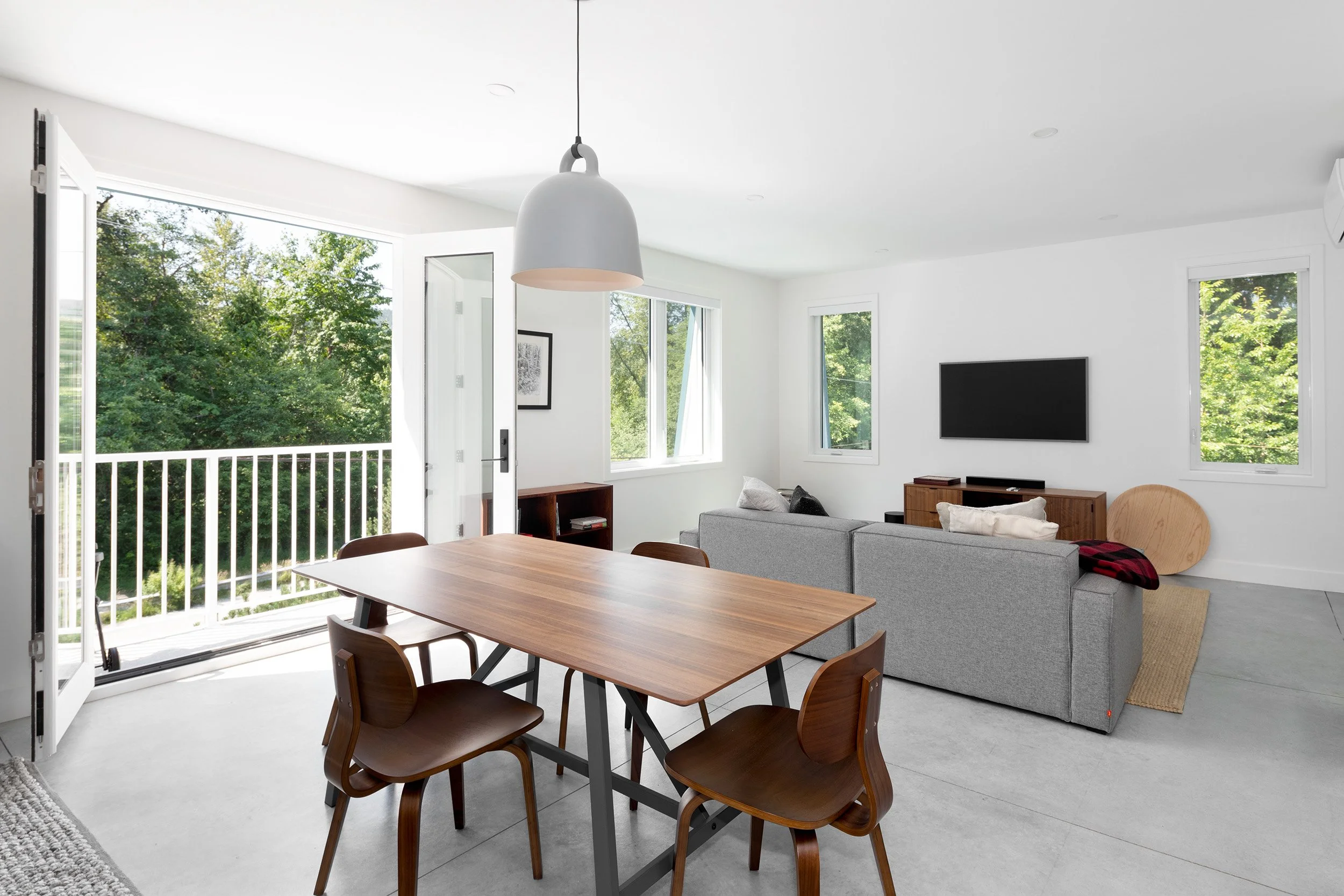 Bright living room with open balcony door, view of green trees outside, modern furniture including a gray sofa, wooden dining table with chairs, wall-mounted TV, large windows, white walls, and a pendant light.