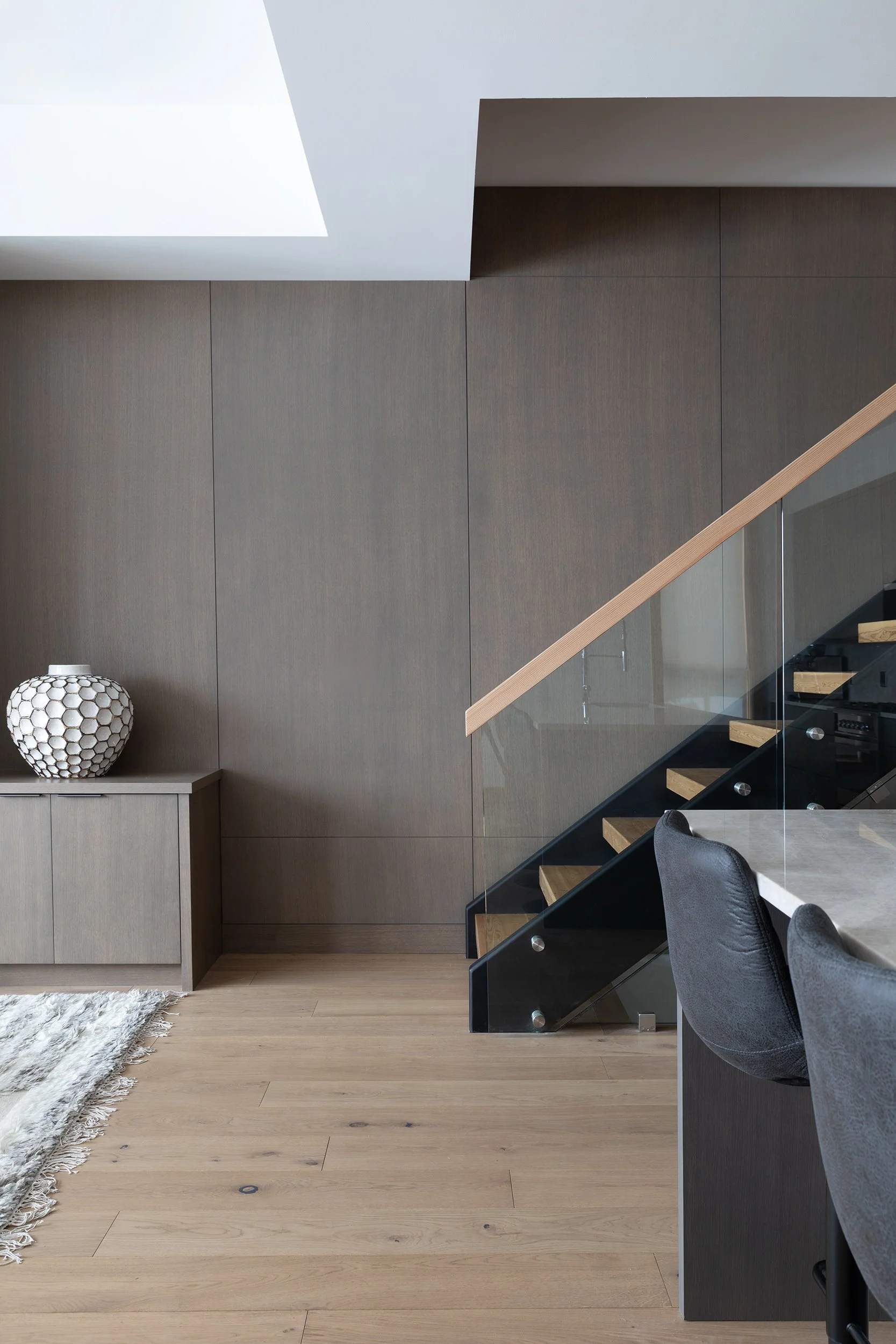 Interior of a modern home featuring a staircase with wood steps and glass railing, beige wood floor, gray upholstered dining chairs, a white sideboard with a geometric-patterned lamp, and a large skylight ceiling.