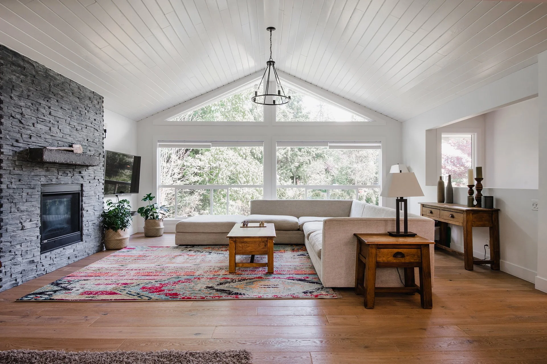 A living room with a vaulted ceiling, a large beige sectional sofa, a wooden side table with a lamp, a colorful area rug, a stone fireplace, a mounted flat-screen TV, and large windows showing trees outside.