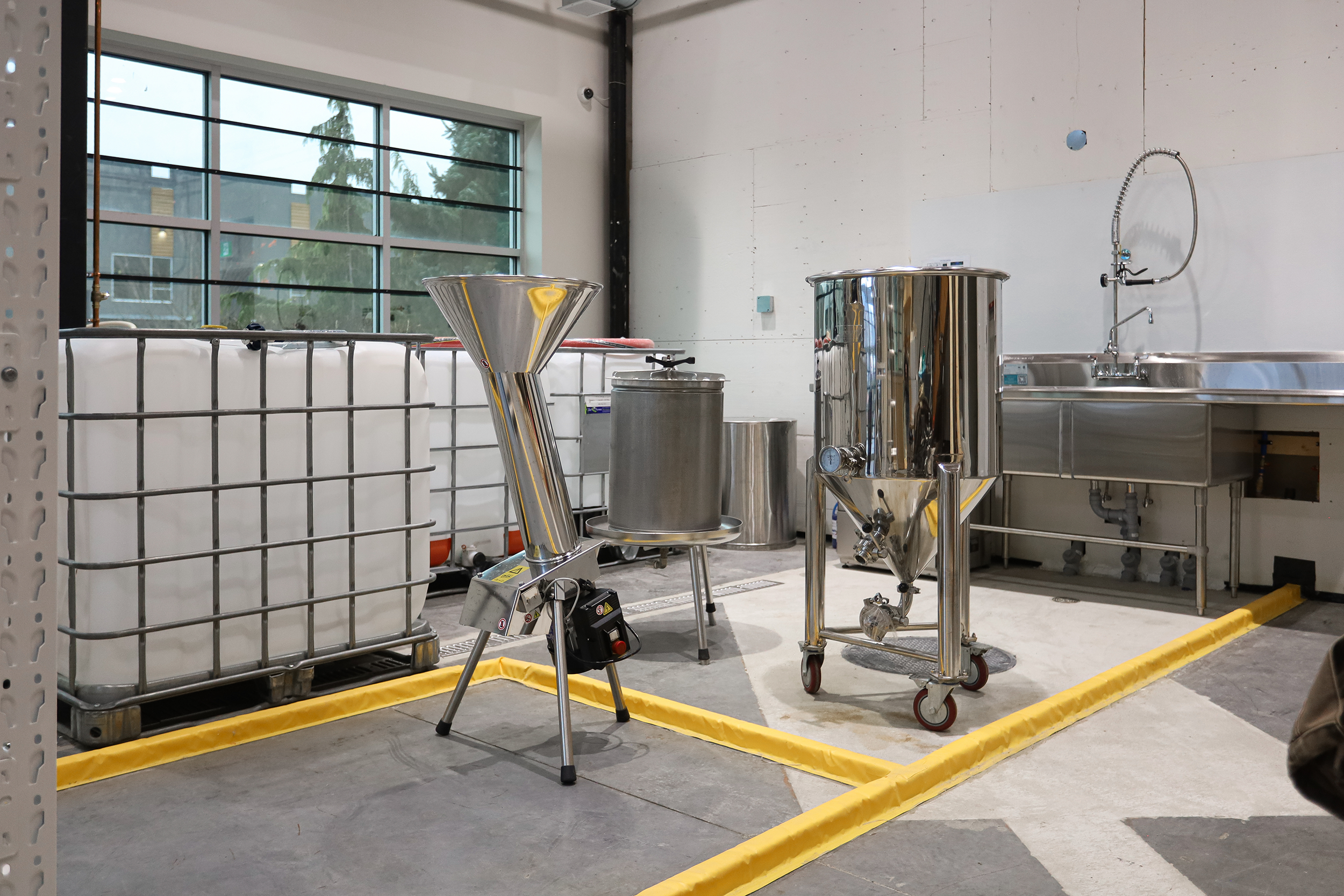 Industrial equipment in a spacious room with large windows, including a stainless steel tank, funnel, and other machinery, with a yellow safety line on the floor.