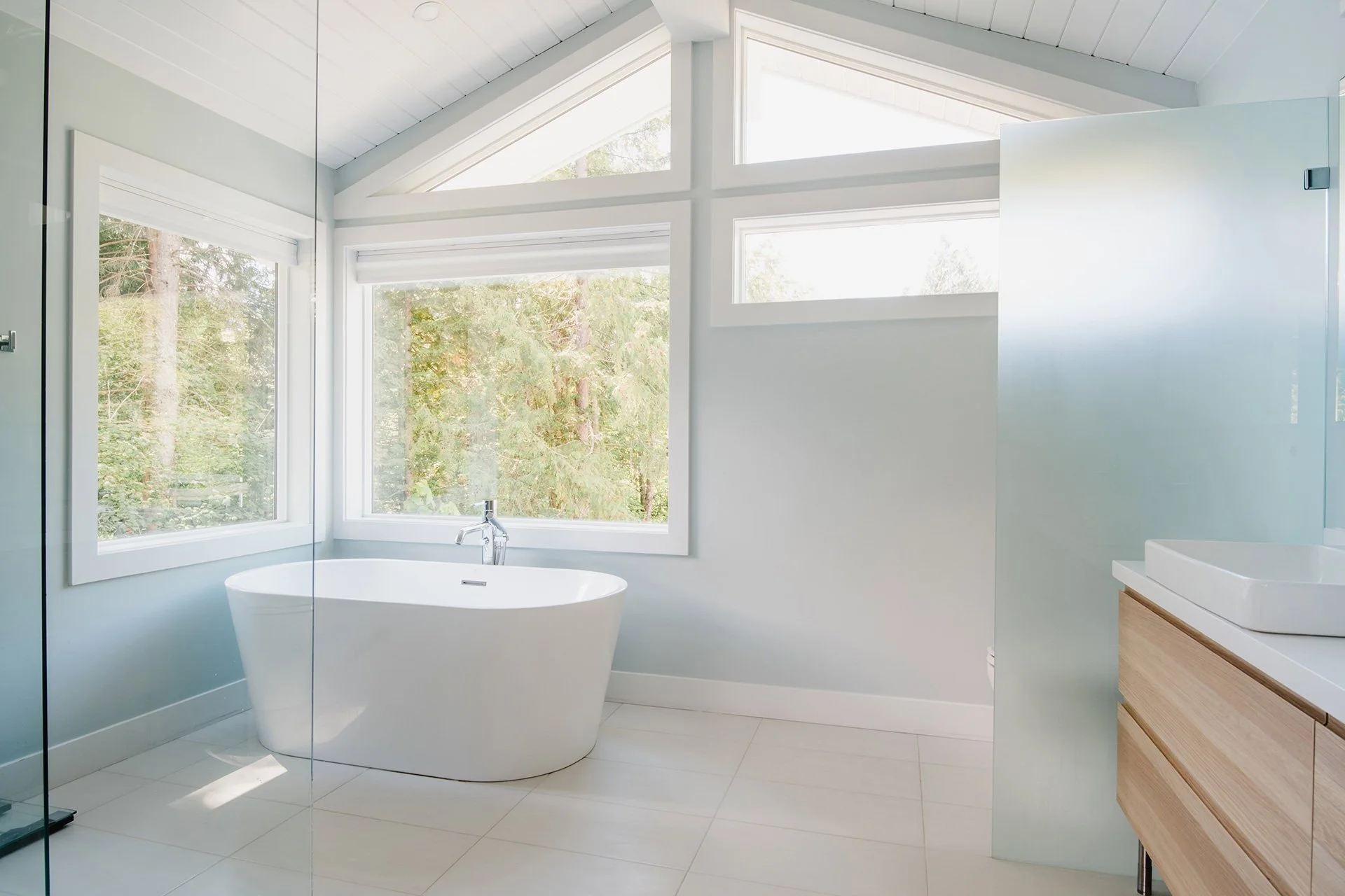 Modern bathroom with large windows overlooking trees, a freestanding bathtub, a white sink with a wooden drawer cabinet, and a frosted glass divider.