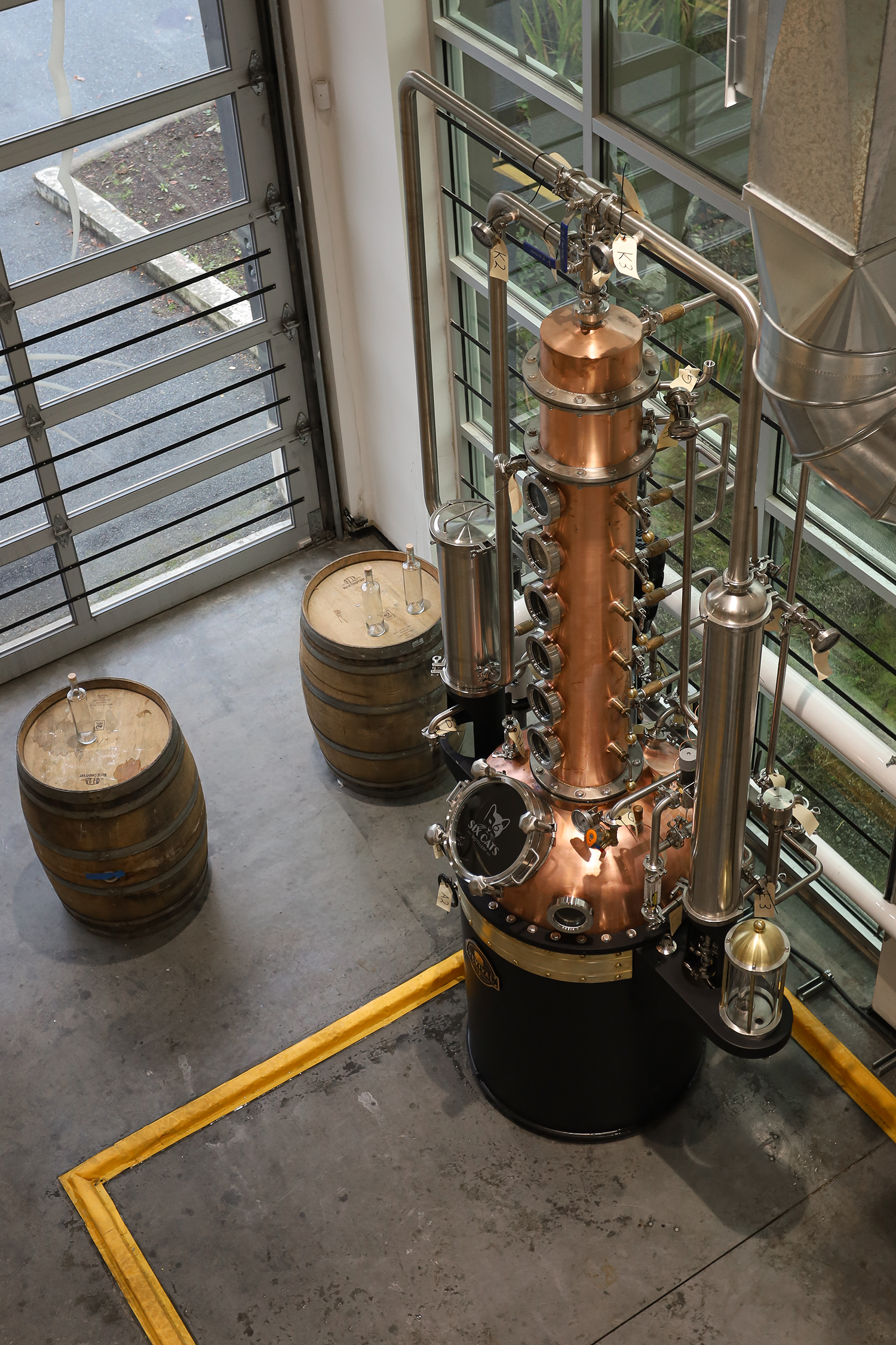 A distillation column and barrels in a distillery, with large windows and an industrial setting.