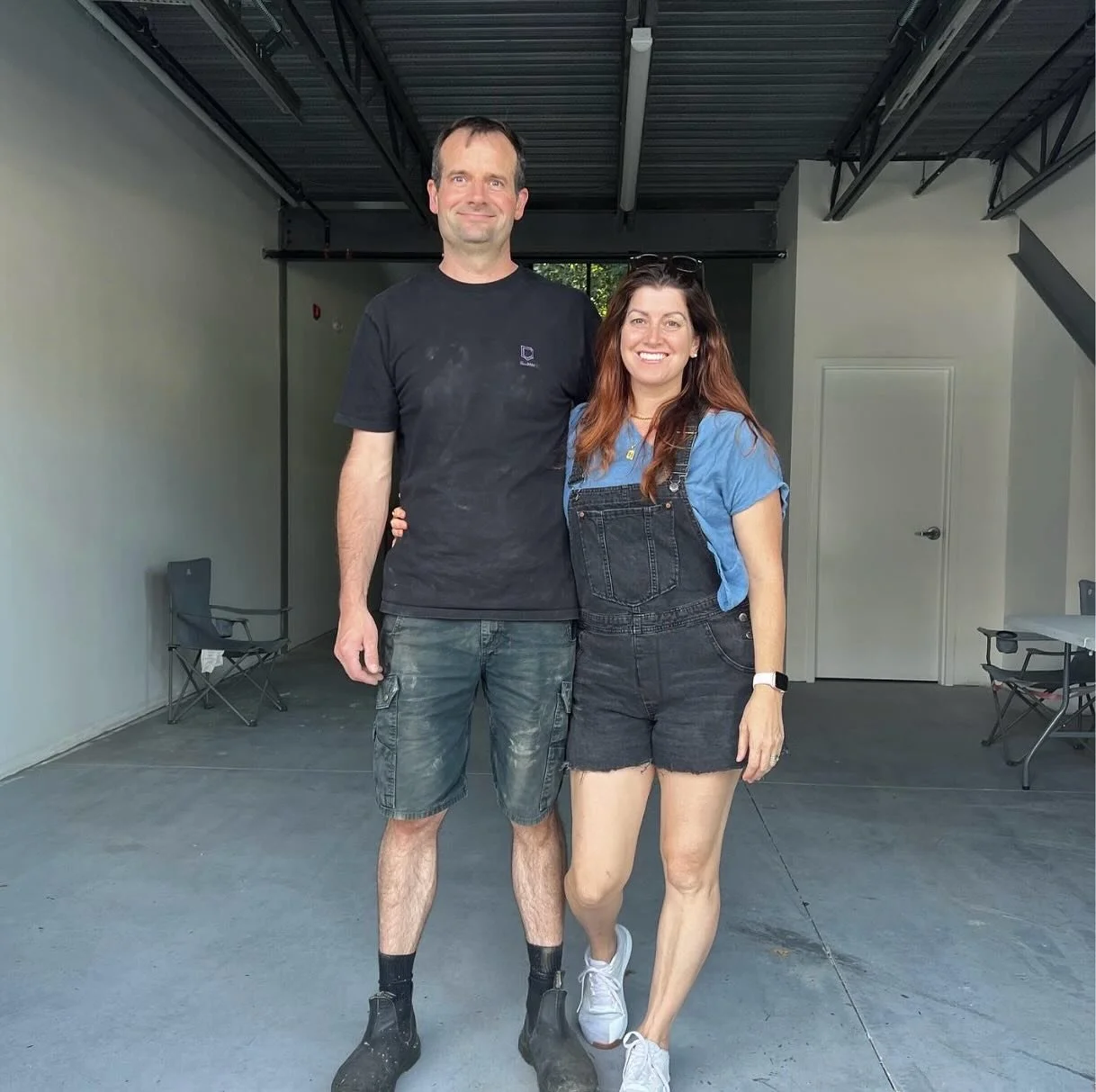 A man and a woman standing together indoors, smiling at the camera. The man is tall, wearing a black t-shirt, denim shorts, and black work boots, and the woman is shorter, with long hair, wearing a blue top, black denim shorts, and white sneakers.
