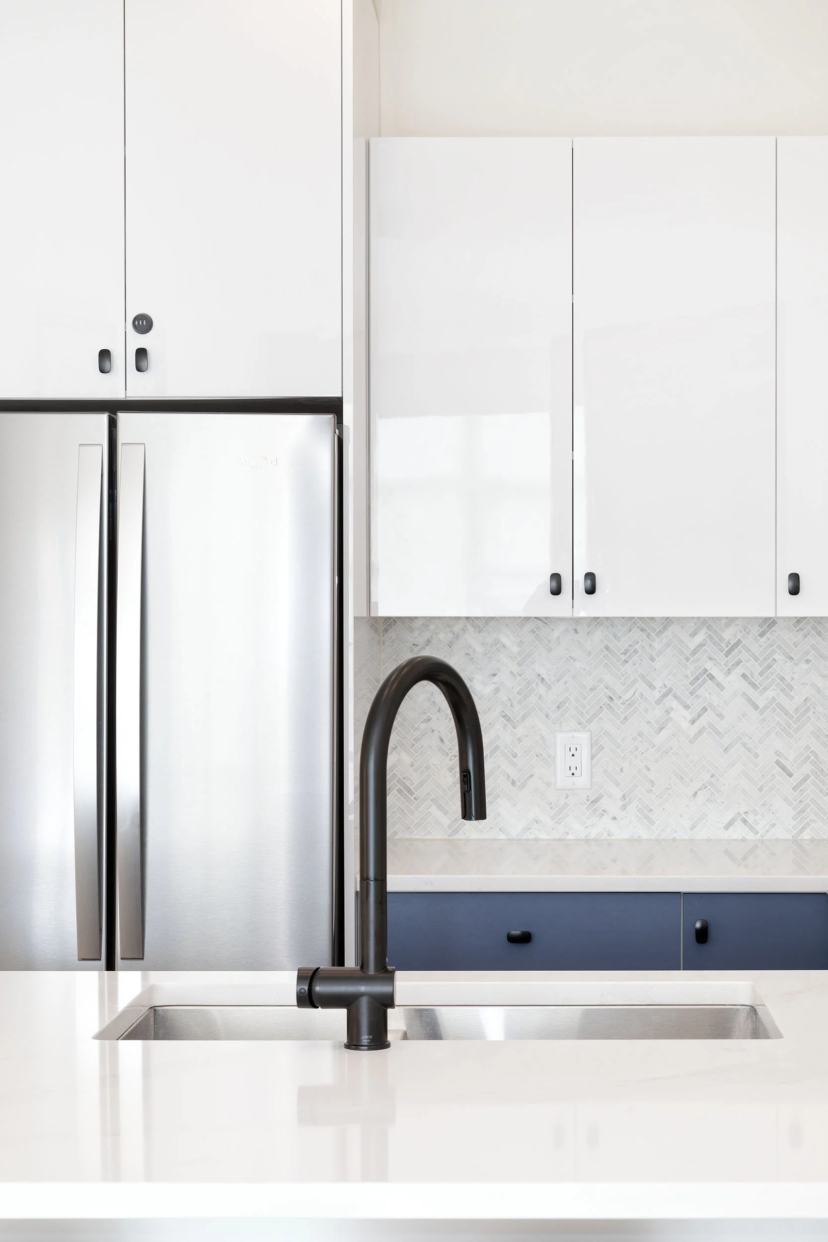 Modern kitchen with white cabinets, stainless steel refrigerator, black faucet over a white sink, and a textured backsplash.