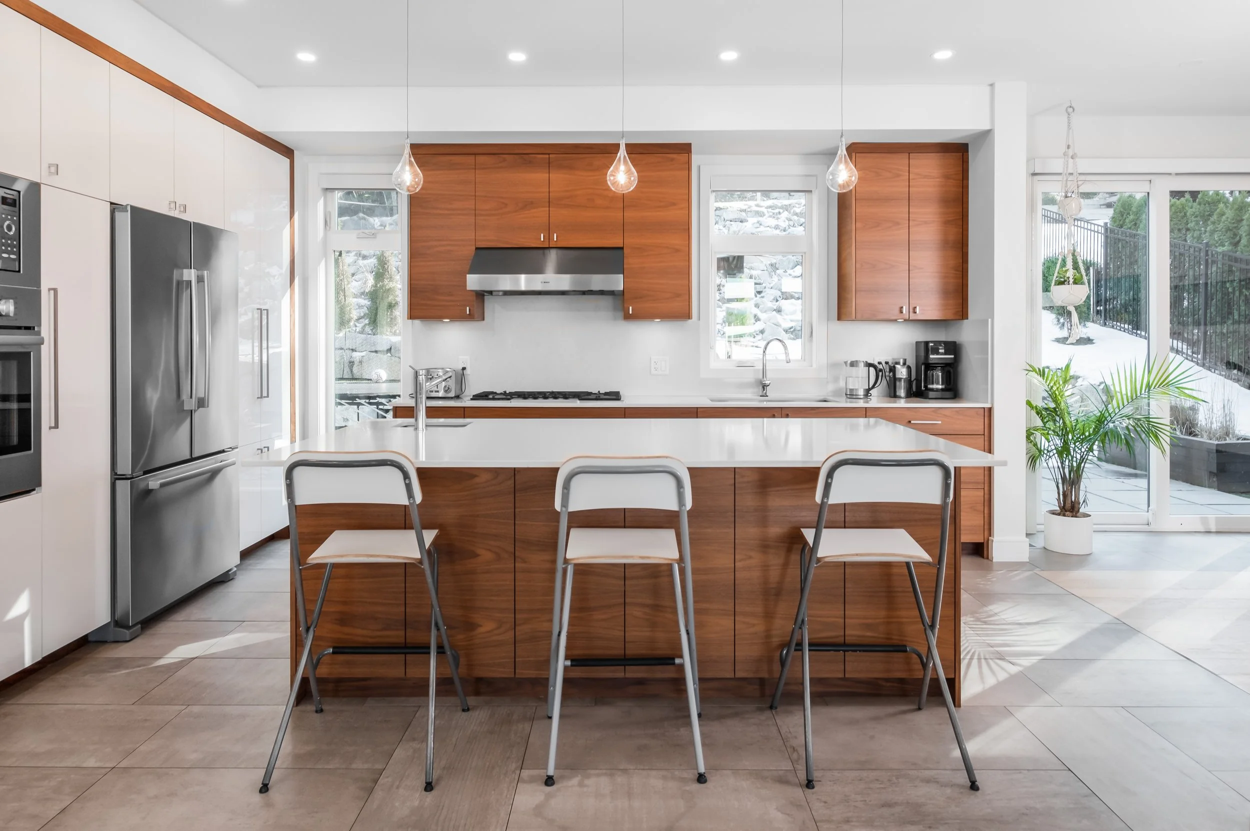 Modern kitchen with white and wood cabinetry, stainless steel appliances, a large central island with three chairs, and large windows showing a snowy outdoor scene with trees and rocks. There are pendant lights hanging over the island and a potted pl