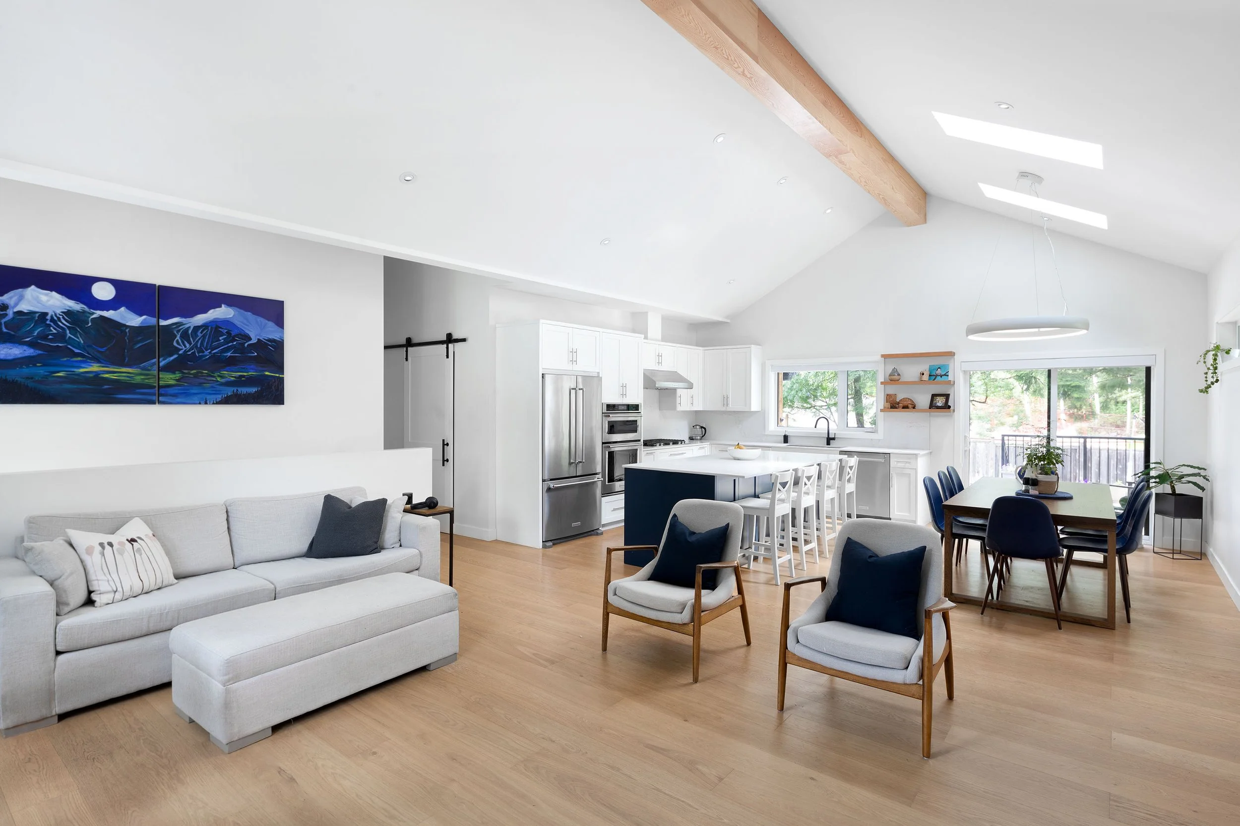 A modern open-concept living space with white walls and wood flooring, featuring a white sofa, a wooden armchair, a dining area with navy chairs, and a kitchen with white cabinets and stainless steel appliances, including a refrigerator and oven.