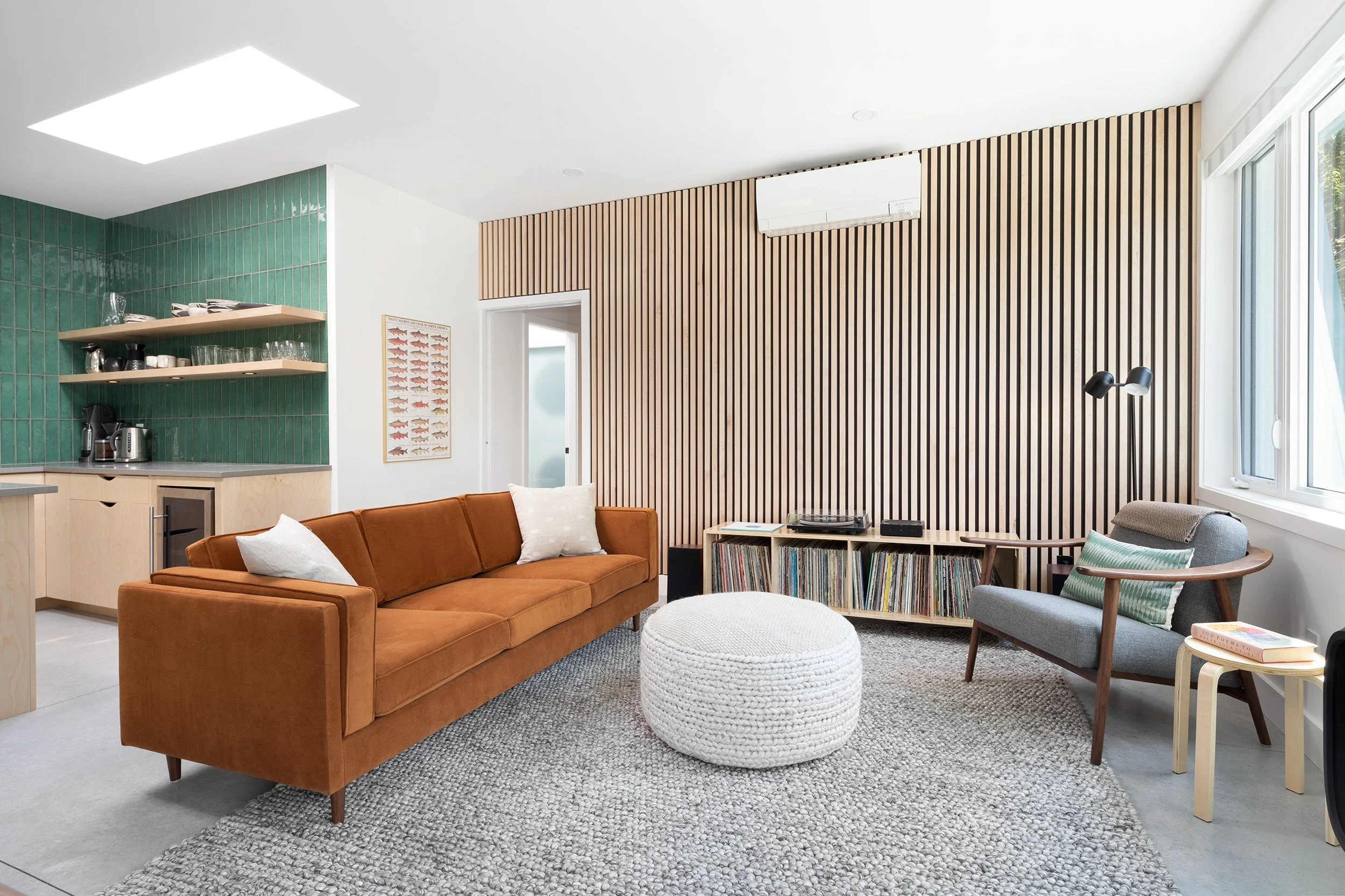 Modern living room with orange sofa, gray armchair, round white pouf, shaggy gray rug, wooden coffee table, bookshelf, and wooden slat wall, illuminated by natural light from large window.