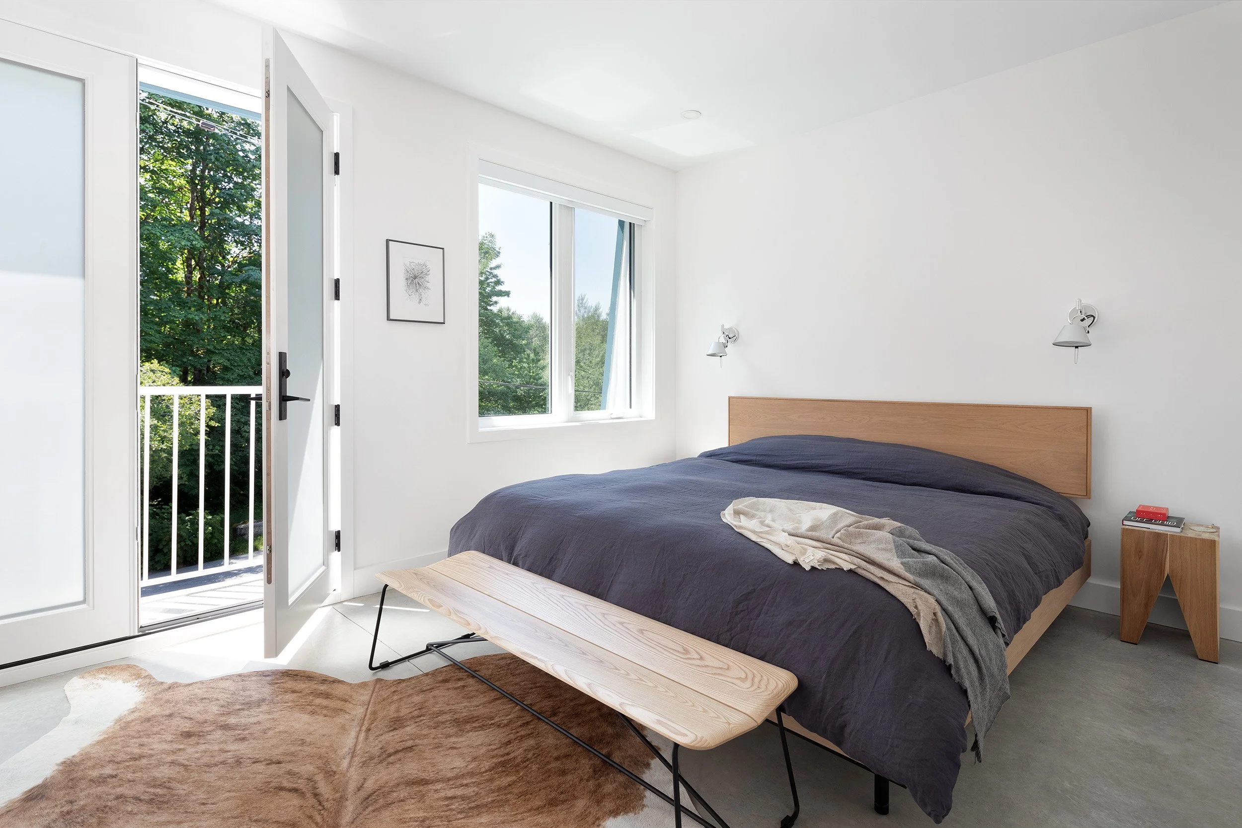 Minimalist bedroom with a large bed, dark bedding, wooden nightstand, wall-mounted lamps, open window with a view of trees, and a glass door leading to a balcony.