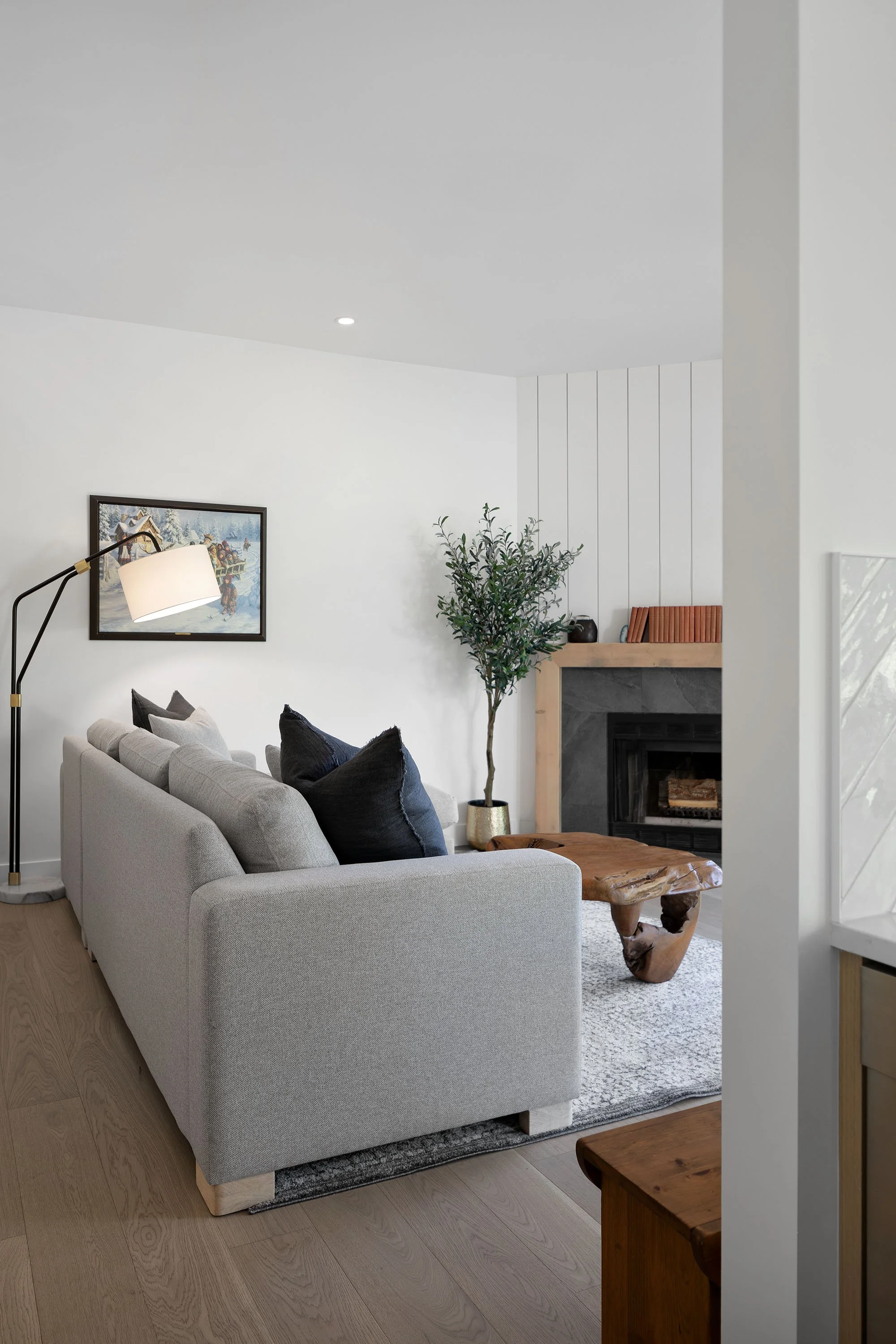 Living room with gray sofa, black and white pillows, wooden coffee table, potted plant, fireplace with wood mantel, and wall art, illuminated by a floor lamp and overhead lighting.