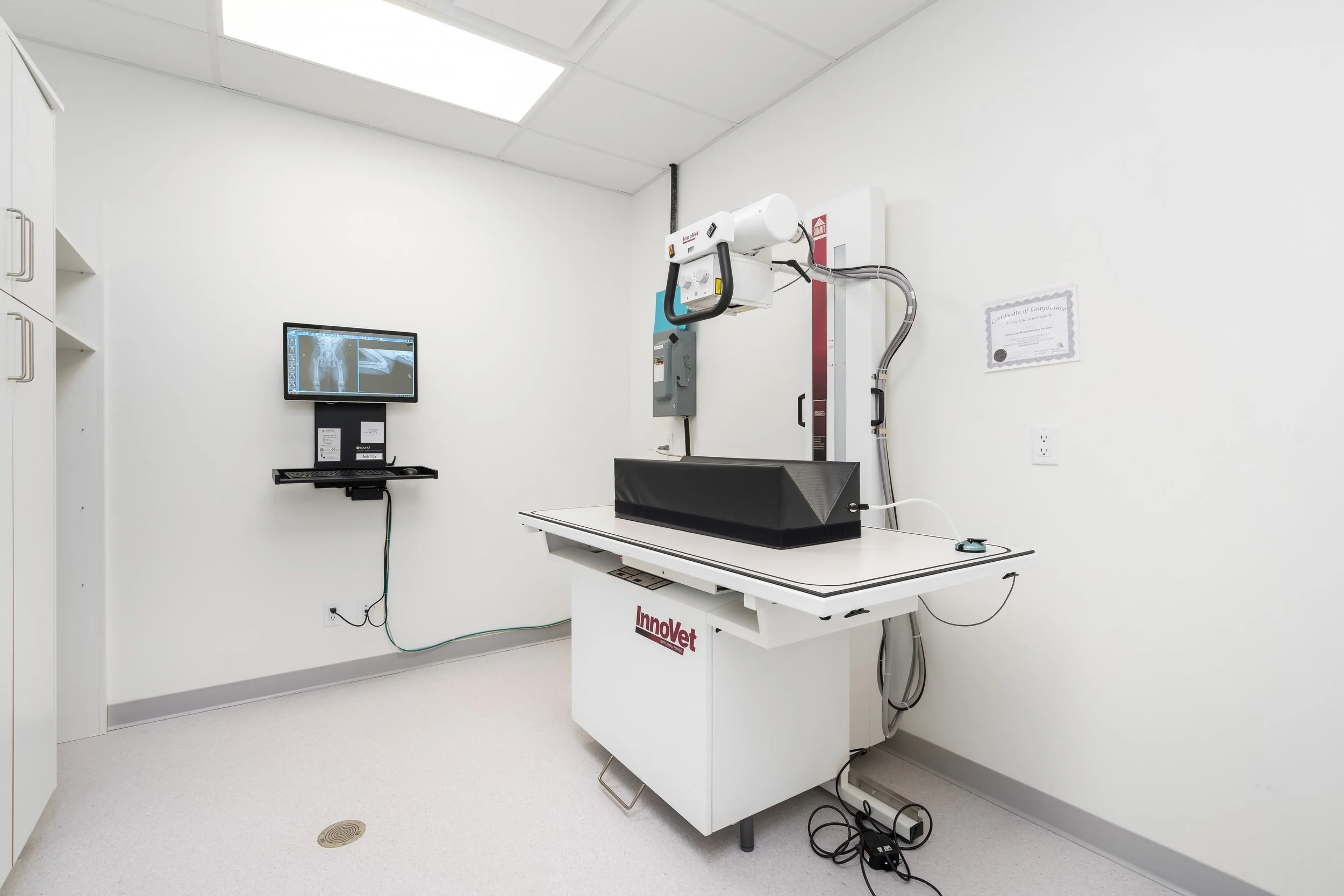 Medical X-ray room with X-ray machine, monitor showing X-ray images, and a white table.