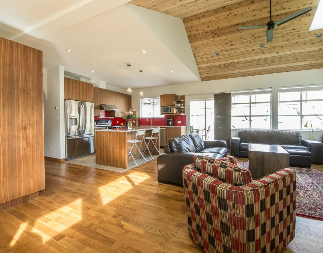 Open-concept living room and kitchen with wood floors, wooden ceiling, large windows, leather sofas, a colorful armchair, and a kitchen island with flowers in a vase.