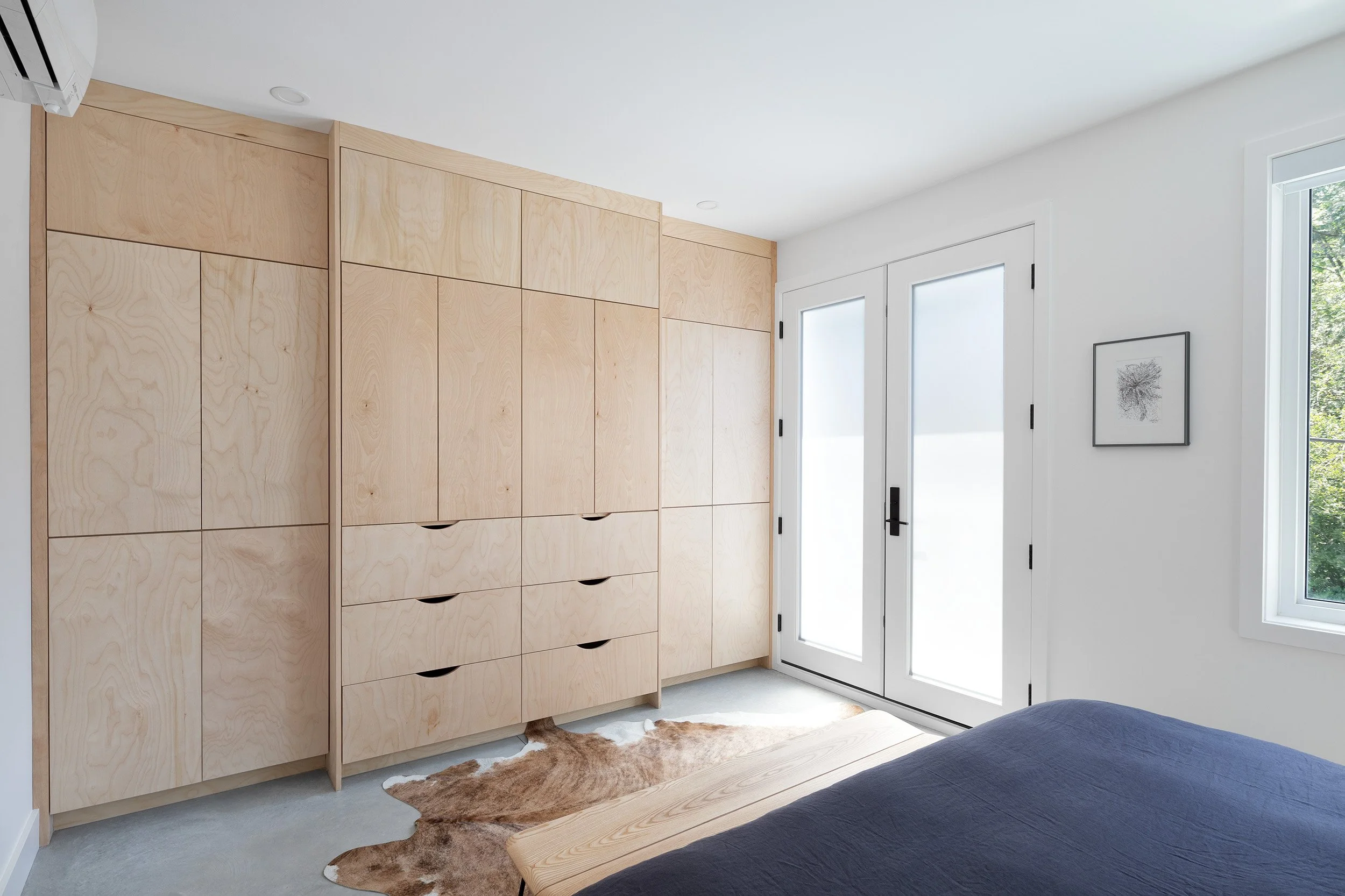 A bedroom corner with light wooden wardrobe, white walls, a window, and a framed artwork. There is a bed with a dark cover and a brown cowhide rug on the floor.