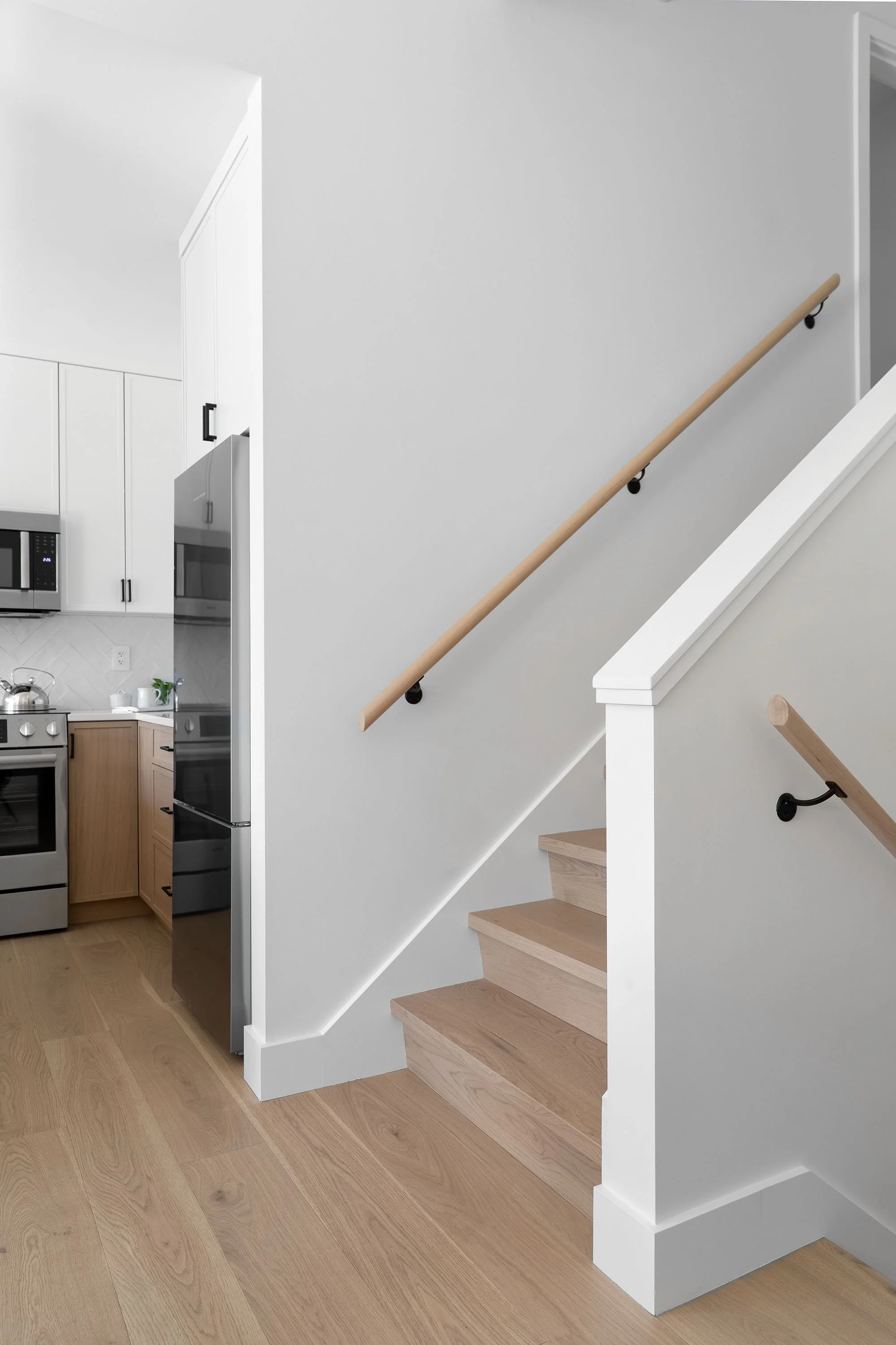 Interior view of a staircase with wooden steps, white banister, and black handrail brackets, adjacent to a kitchen with white cabinets, stainless steel appliances, and light wood flooring.