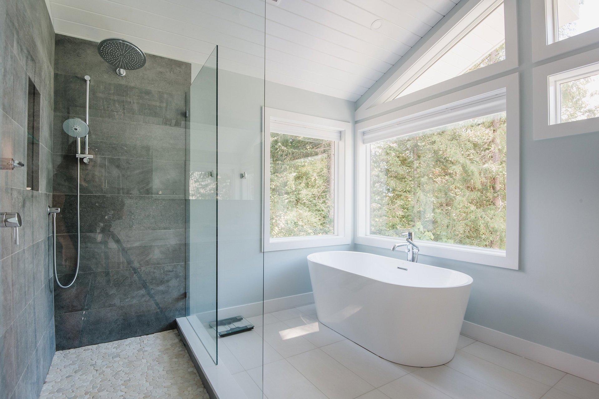 Modern bathroom featuring a walk-in shower with dark gray tiles and glass enclosure, and a freestanding bathtub beneath large corner windows with a view of trees outside.