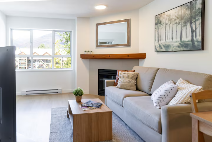 Living room with a beige sofa, pillows, a wooden coffee table with magazines and a potted plant, a fireplace, large window, and wall art of trees.