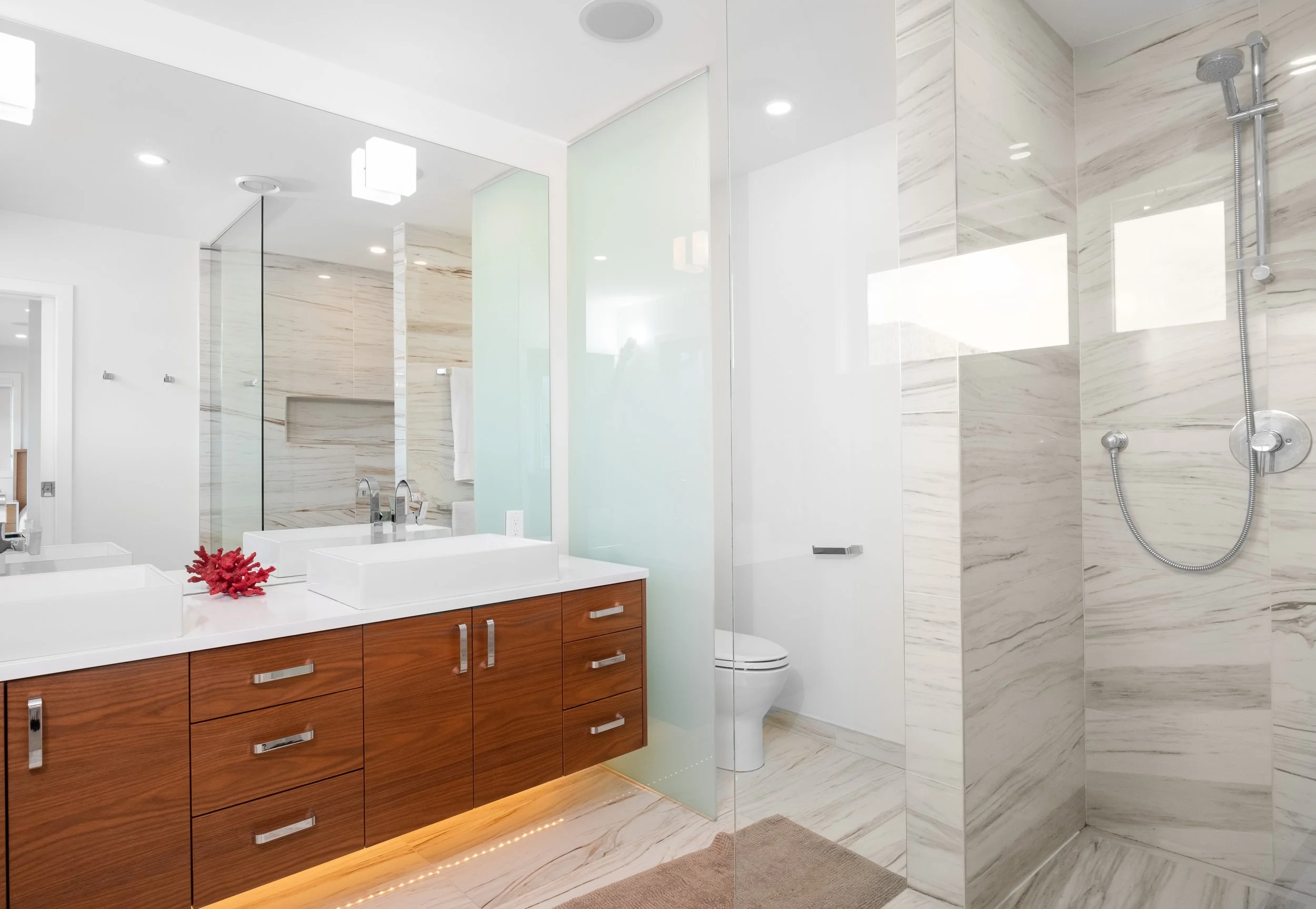 Modern bathroom with wood vanity, white sinks, large mirror, marble walls and floor, glass shower enclosure, with shower fixtures and a robe hook visible.