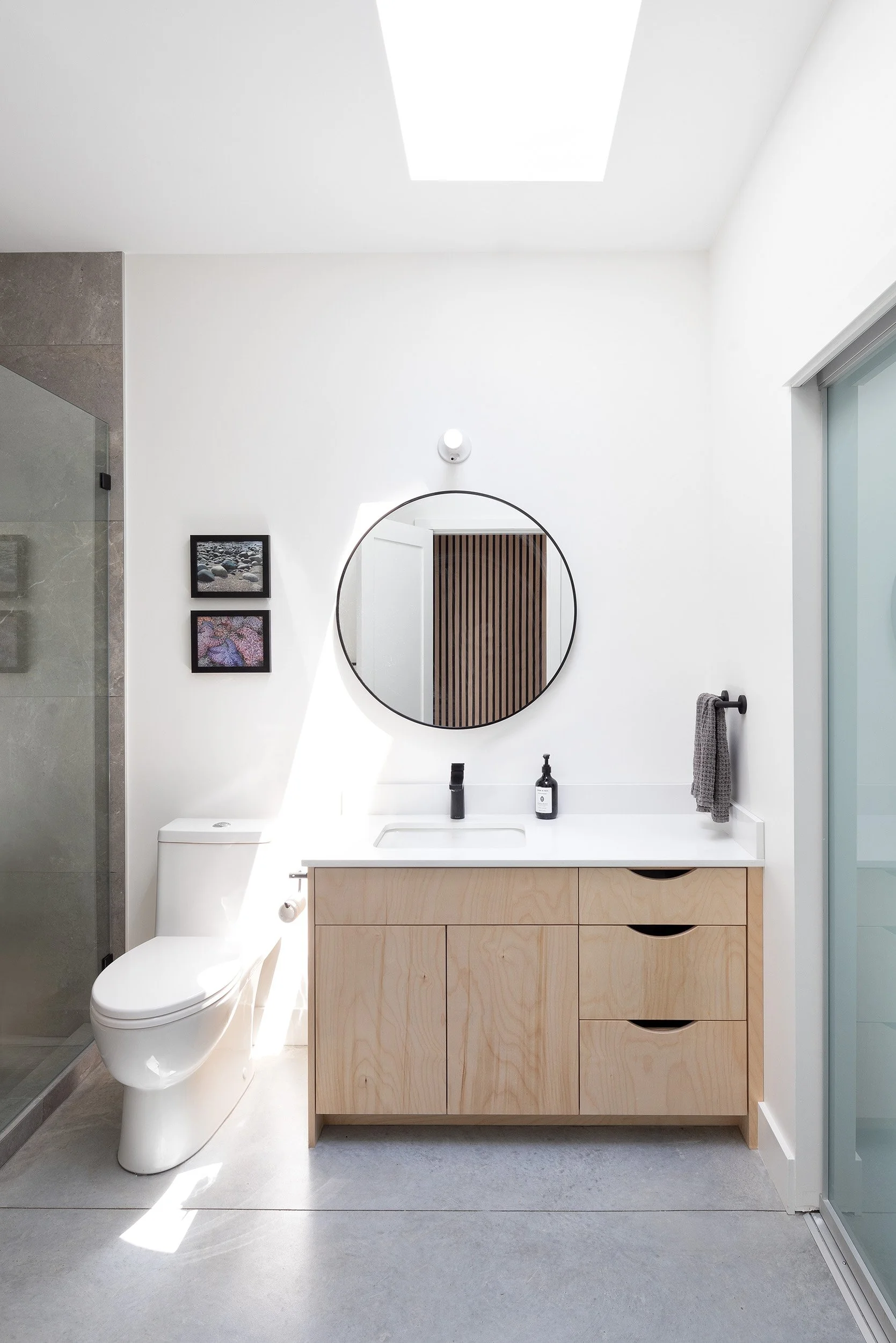 Modern bathroom with a white toilet, wooden vanity with storage drawers, a round mirror, and framed pictures on the wall. Natural light from a skylight illuminates the space.