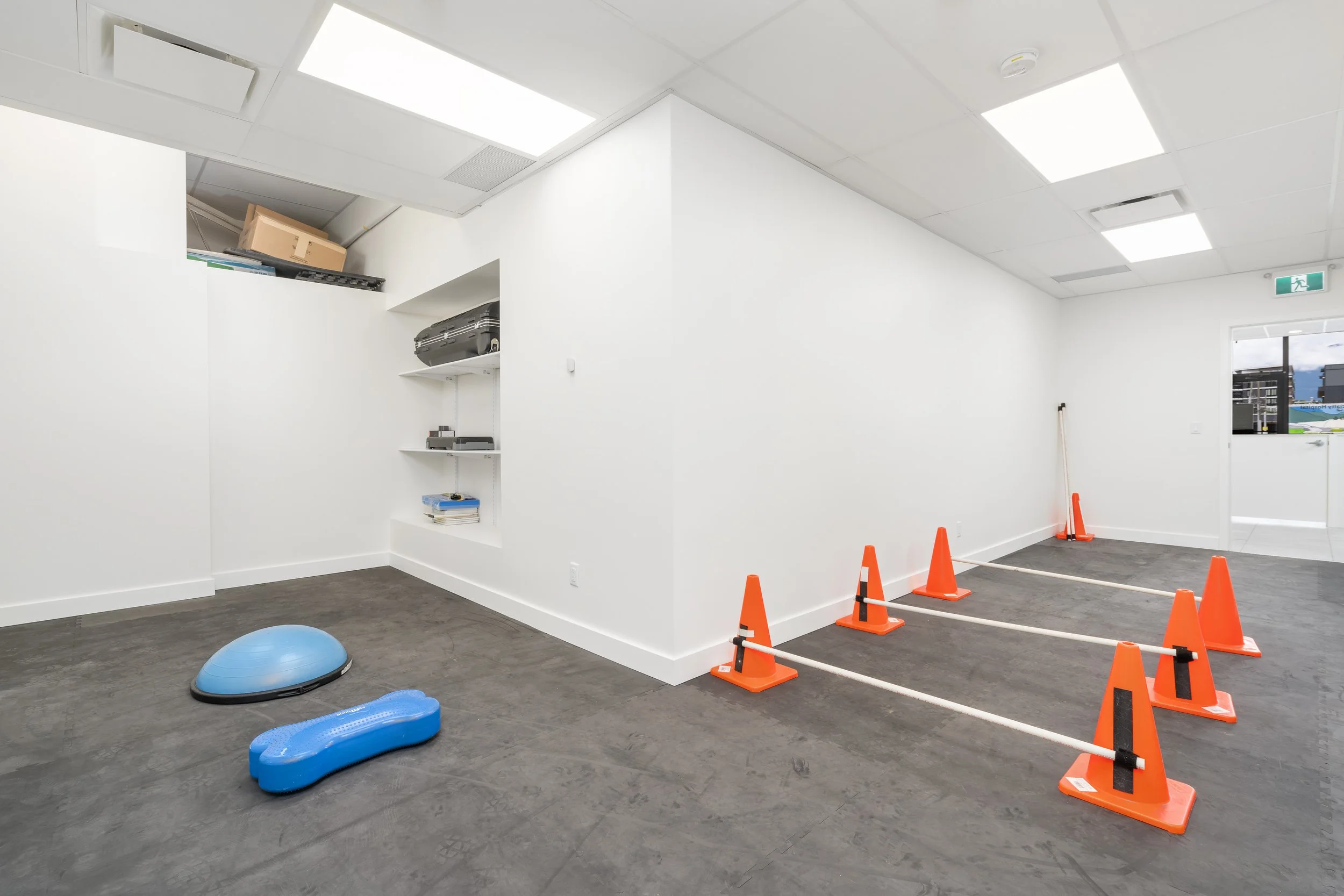Empty room with white walls and ceiling, orange cones and barrier tape on the dark floor, a blue exercise ball, and shelves with boxes and equipment.