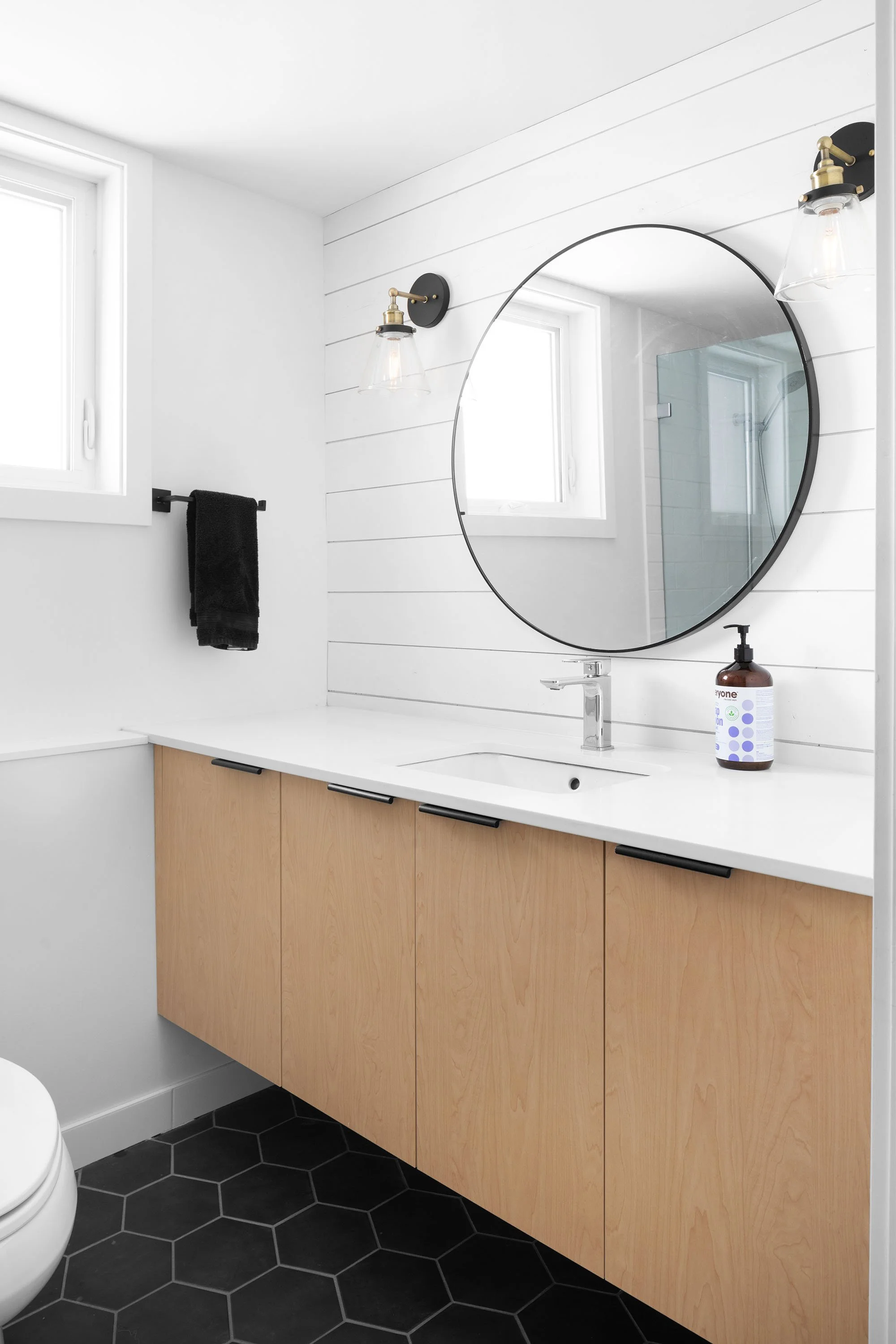 Modern bathroom vanity with a round mirror, dual wall sconces, a black towel, and a soap dispenser on a white countertop with light wood cabinets.