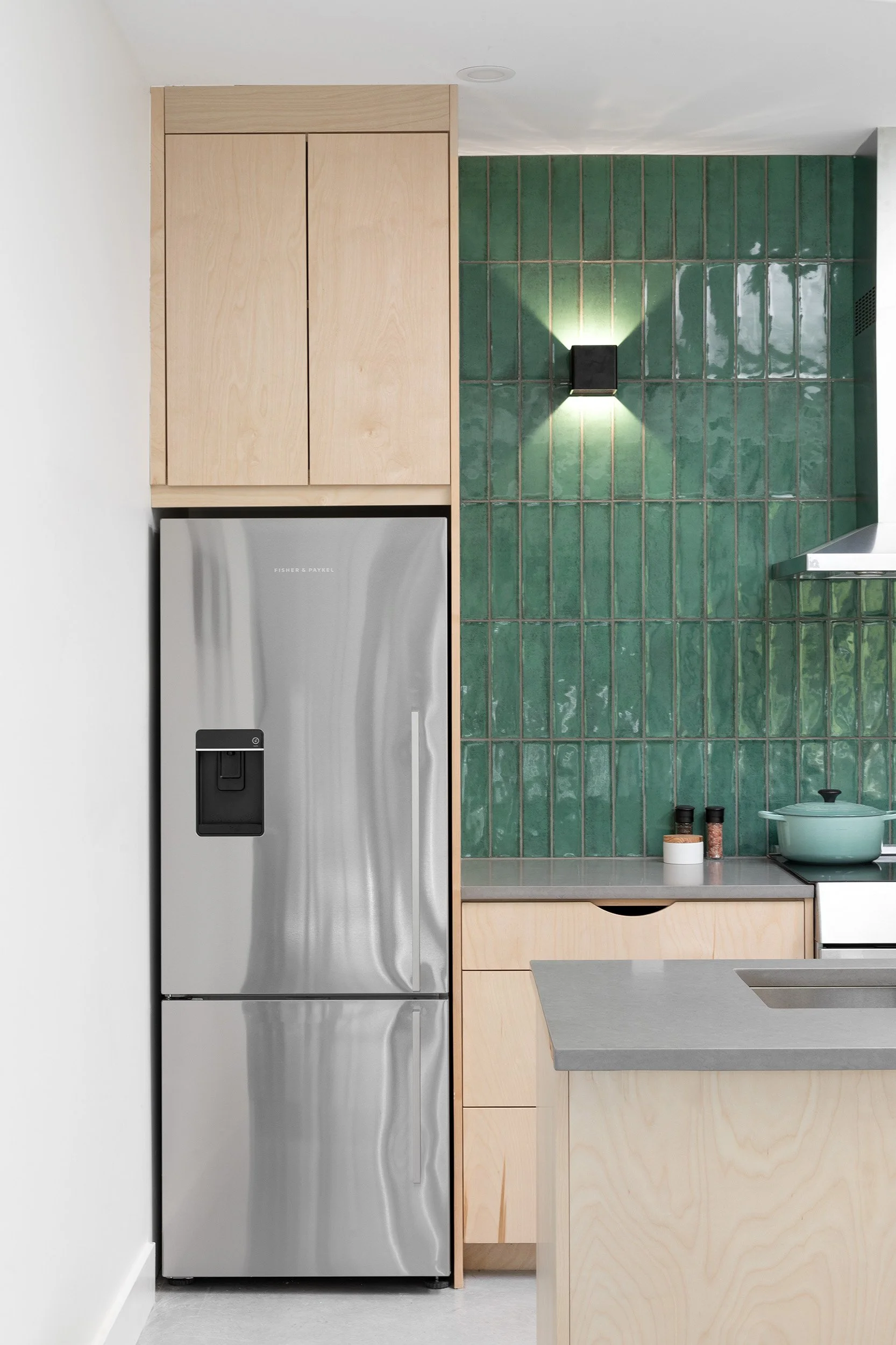 A kitchen with a stainless steel refrigerator, light wooden cabinetry, green tiled backsplash, and a countertop with a teal pot and spice jars.