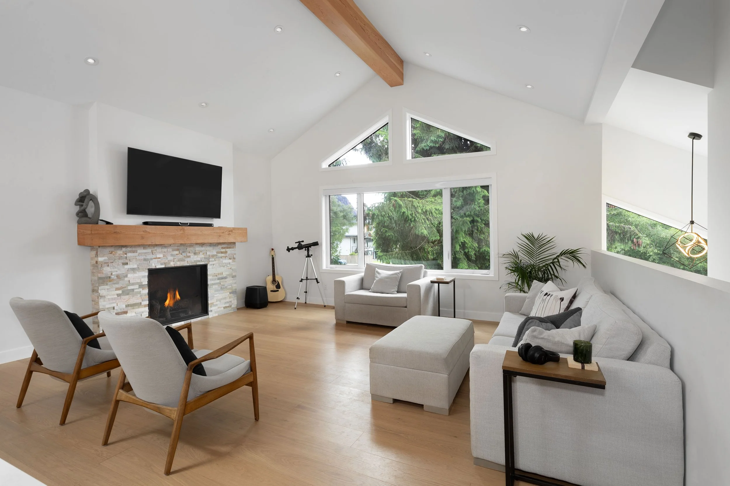 A bright, modern living room with white walls and a vaulted ceiling featuring wooden beams. There is a stone fireplace with a wooden mantel, a wall-mounted flat-screen TV, and a small sculpture on the mantel. Large windows show green trees outside. The room has a white sofa, armchairs, a guitar, a telescope, and a unique pendant light fixture. Hardwood flooring completes the cozy and inviting space.