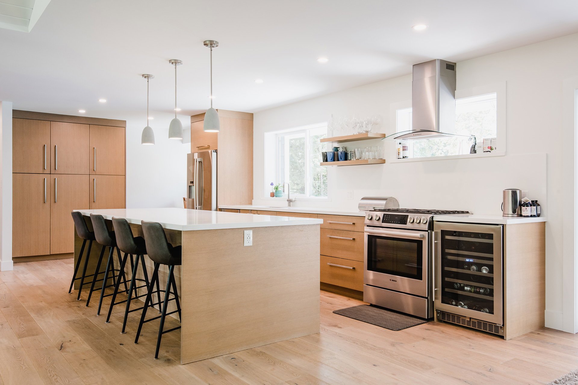 Modern kitchen with wooden cabinets, stainless steel appliances, a white island with black chairs, and natural light from large windows.