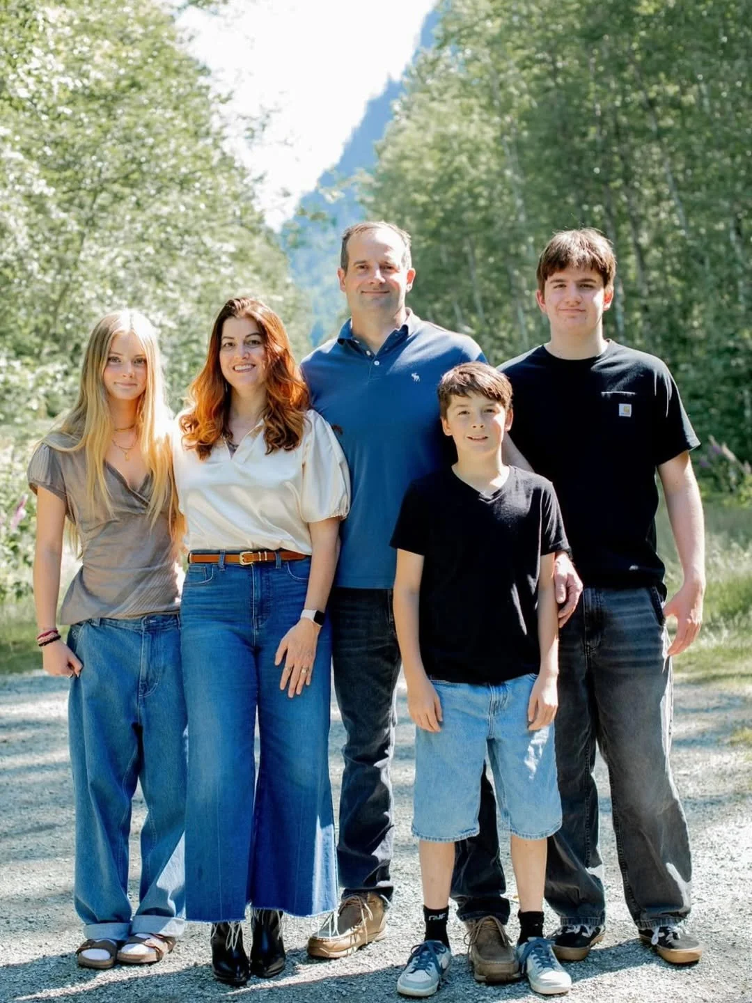 Family of five outdoors on a sunny day, standing on a dirt path surrounded by trees with mountains in the background.