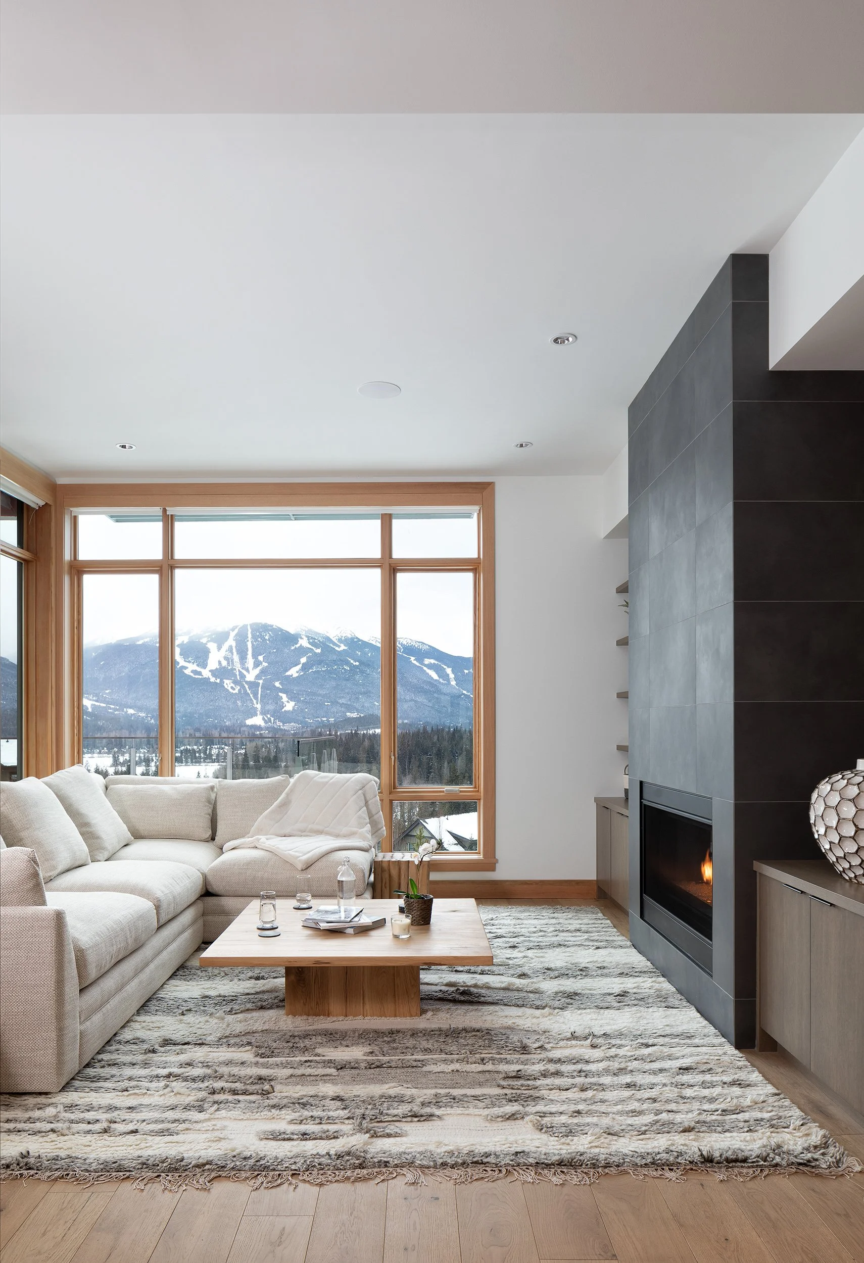 Living room with large window showing snowy mountain view, beige couch with cushions, wooden coffee table with decor, stone fireplace, and patterned rug.