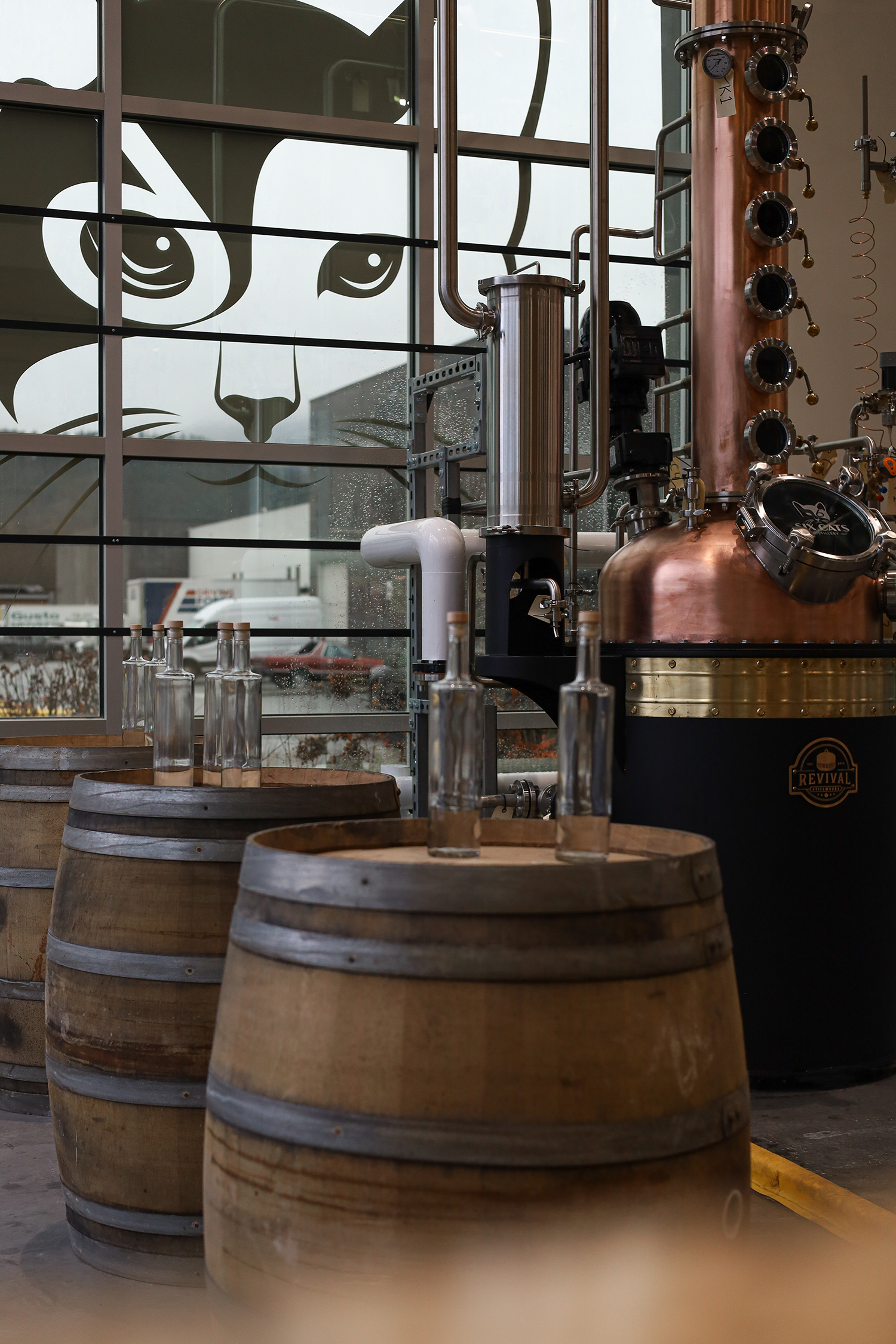 View of three wooden barrels used for aging beverages, positioned in front of a large window with a lion face logo decal. Inside, there is brewing or distillation equipment made of copper and stainless steel.
